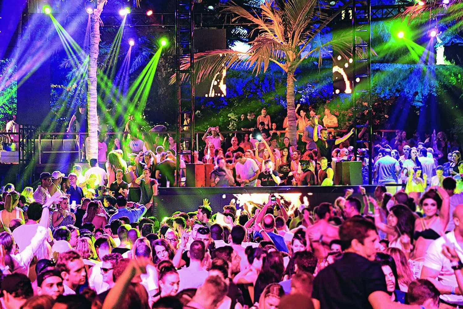 Crowded outdoor nightclub scene with people dancing under colorful stage lights and palm trees.
