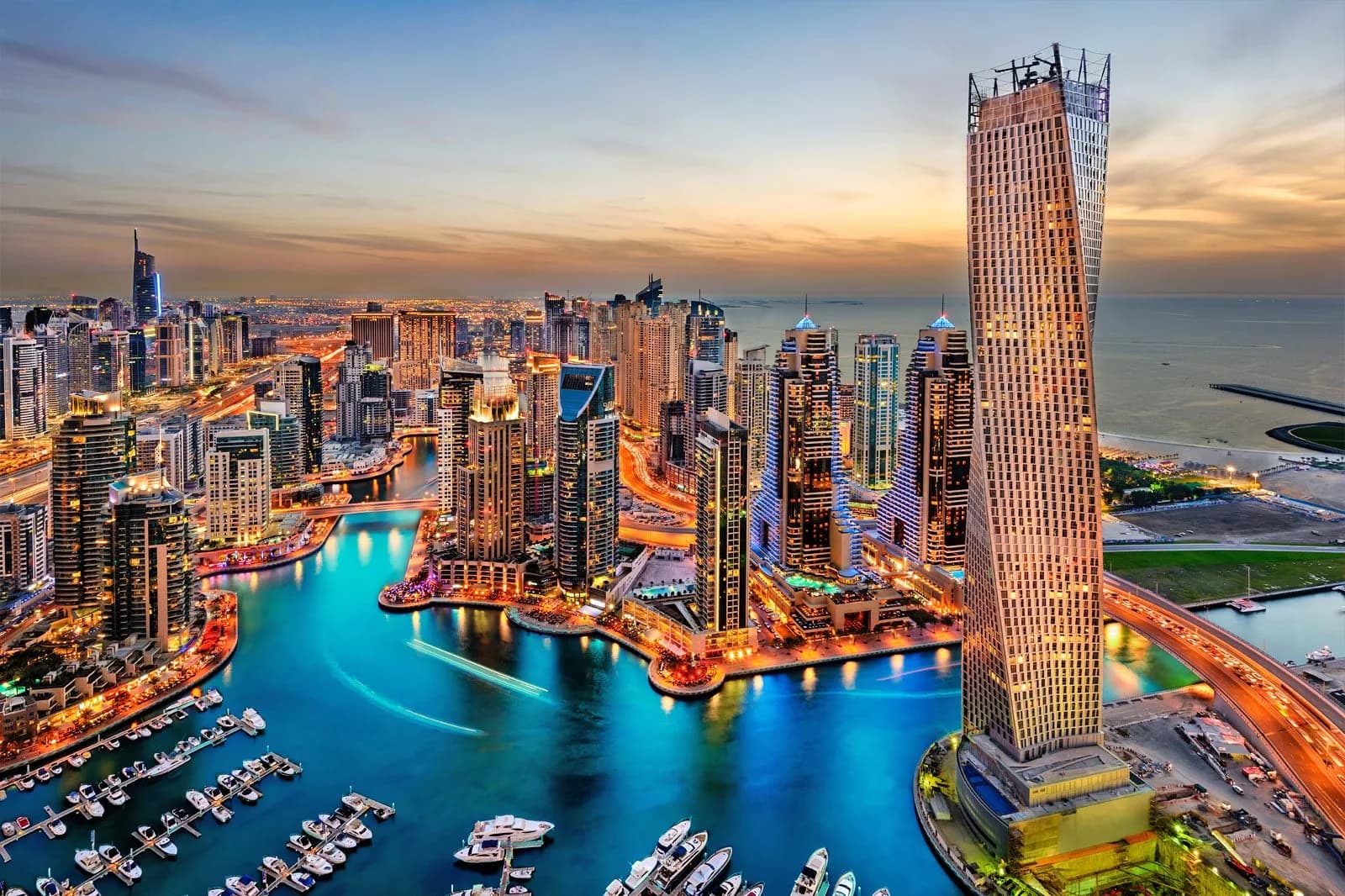 Aerial view of a vibrant city skyline with a prominent twisted skyscraper near the waterfront, surrounded by numerous high-rise buildings and illuminated at twilight, with a marina filled with boats in the foreground.