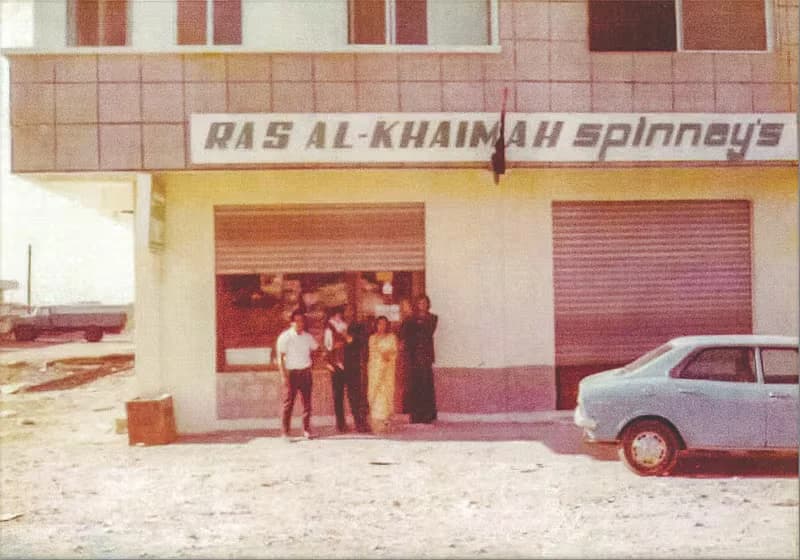 A vintage photograph of a building with the sign "Ras Al-Khaimah Spinneys". Four people stand in front of the building entrance, and a light blue car is parked nearby. The setting appears to be an arid, undeveloped area.