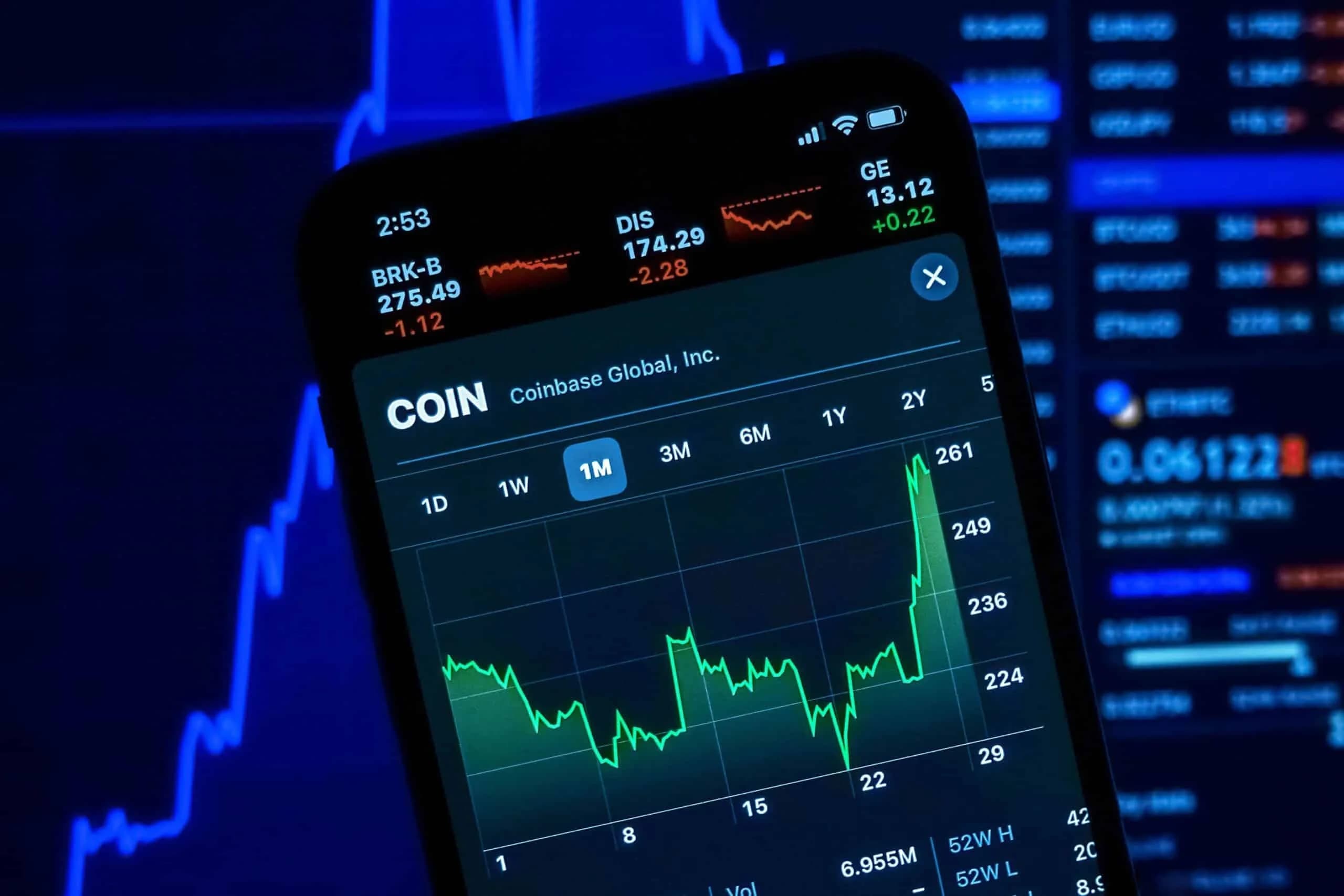 A smartphone displaying a stock chart for Coinbase Global, Inc., with a graph showing price fluctuations over a one-month period. The background shows blurred financial data and stock prices on a screen.