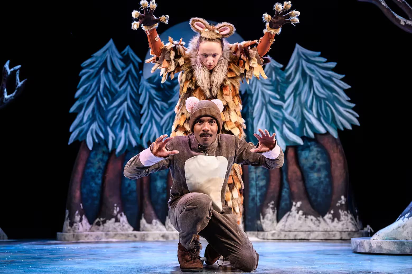 Two actors in animal costumes performing on stage with a forest backdrop. The actor in front is wearing a brown costume with ears resembling a small creature, while the actor behind is dressed in a larger, brown and tan costume with large ears and is standing with raised arms.