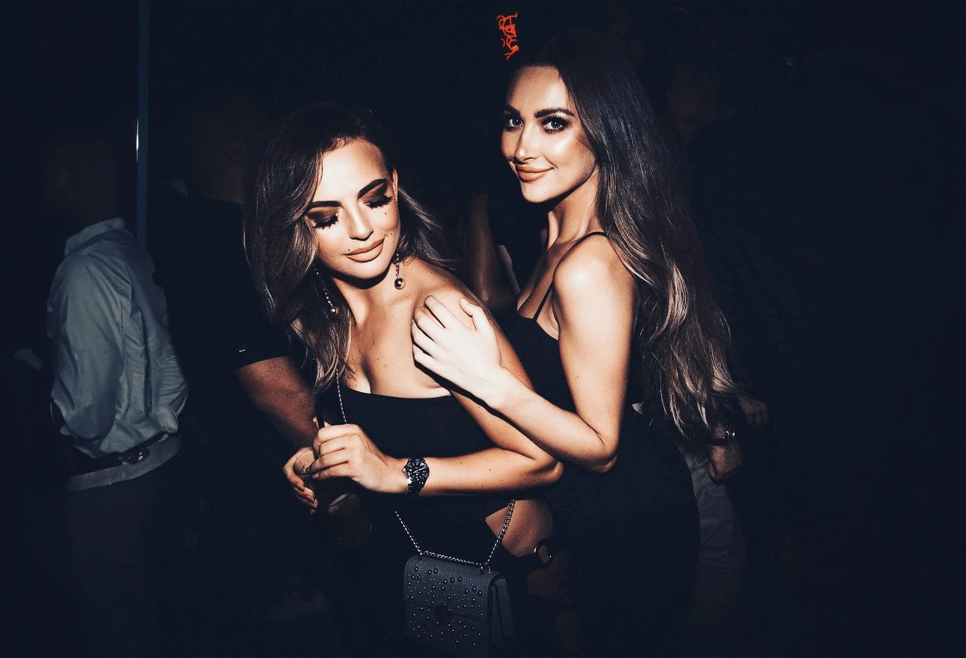 Two women in black dresses are smiling and posing together in a dimly lit environment.