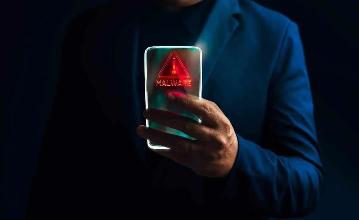 A person in a dark suit holds a smartphone displaying a glowing red malware warning with an exclamation mark inside a triangle.