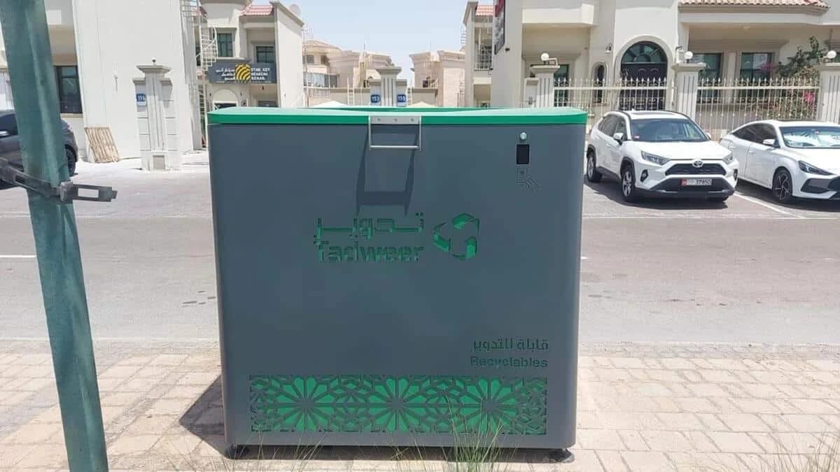 A large gray recycling bin with a green top, labeled "Tadweer Recyclables" in both Arabic and English, situated on a street with parked cars and buildings in the background.