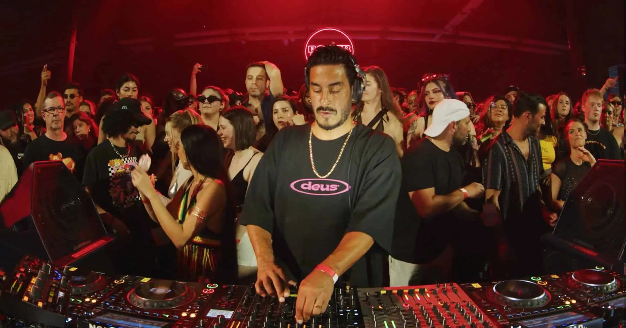 A DJ performing at a crowded event, surrounded by people under red lighting. The DJ is wearing a black shirt with the word 'deus' on it and has headphones around his neck. A DJ console with controls and turntables is visible in the foreground.