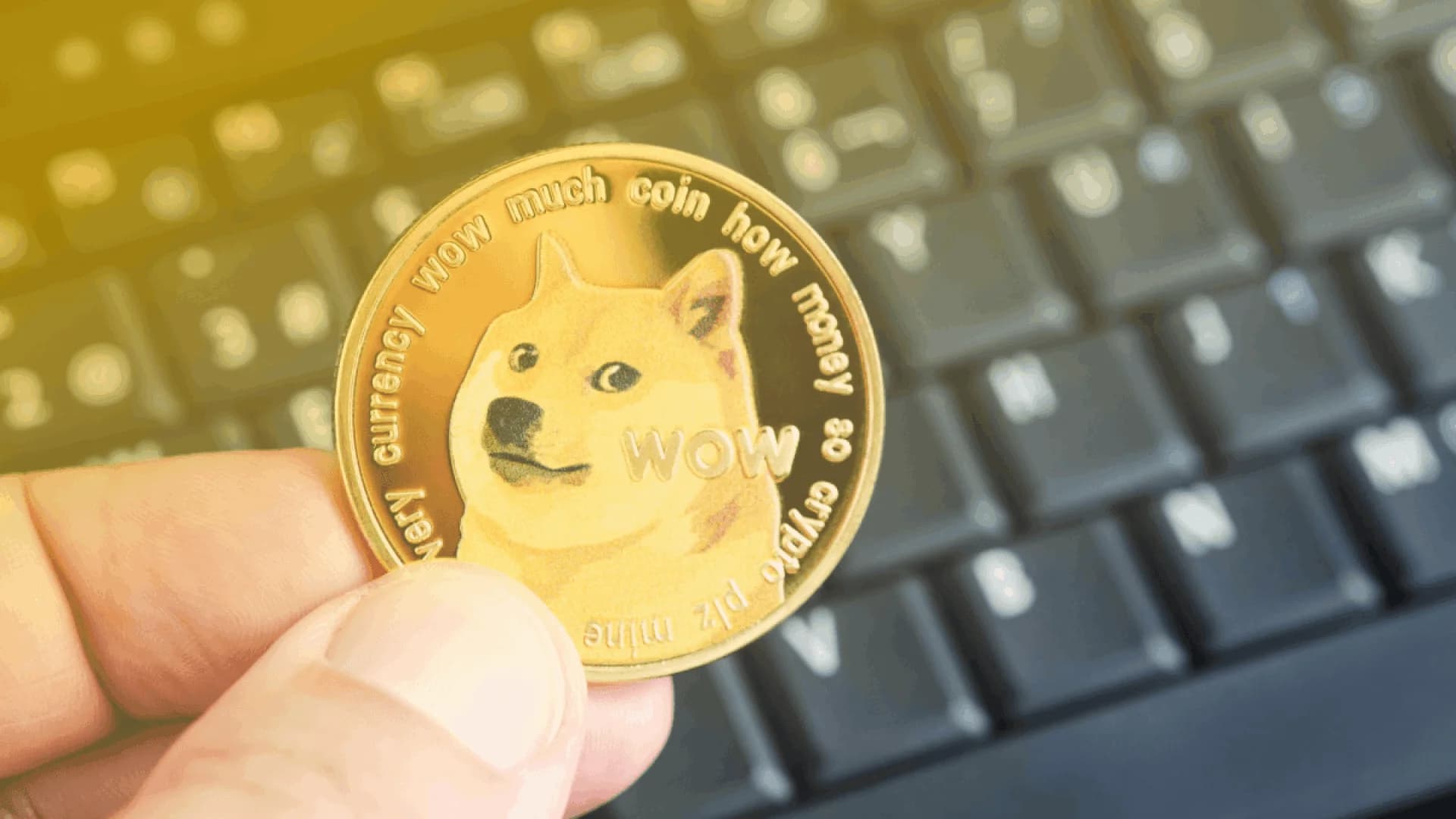 A person holding a Dogecoin, featuring the Shiba Inu meme dog, in front of a computer keyboard.