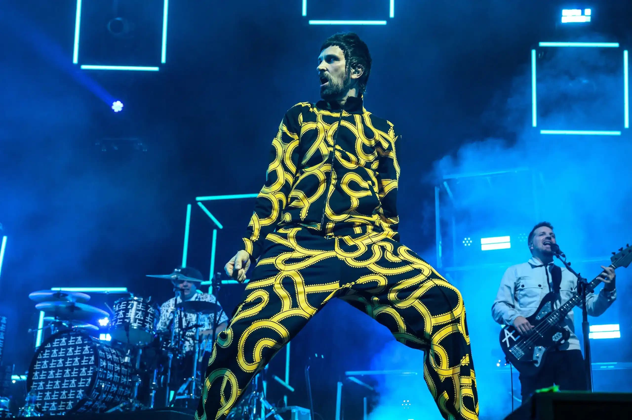 A performer in a vibrant, patterned yellow and black outfit sings energetically on stage, with a drummer and bassist in the background under blue stage lighting.
