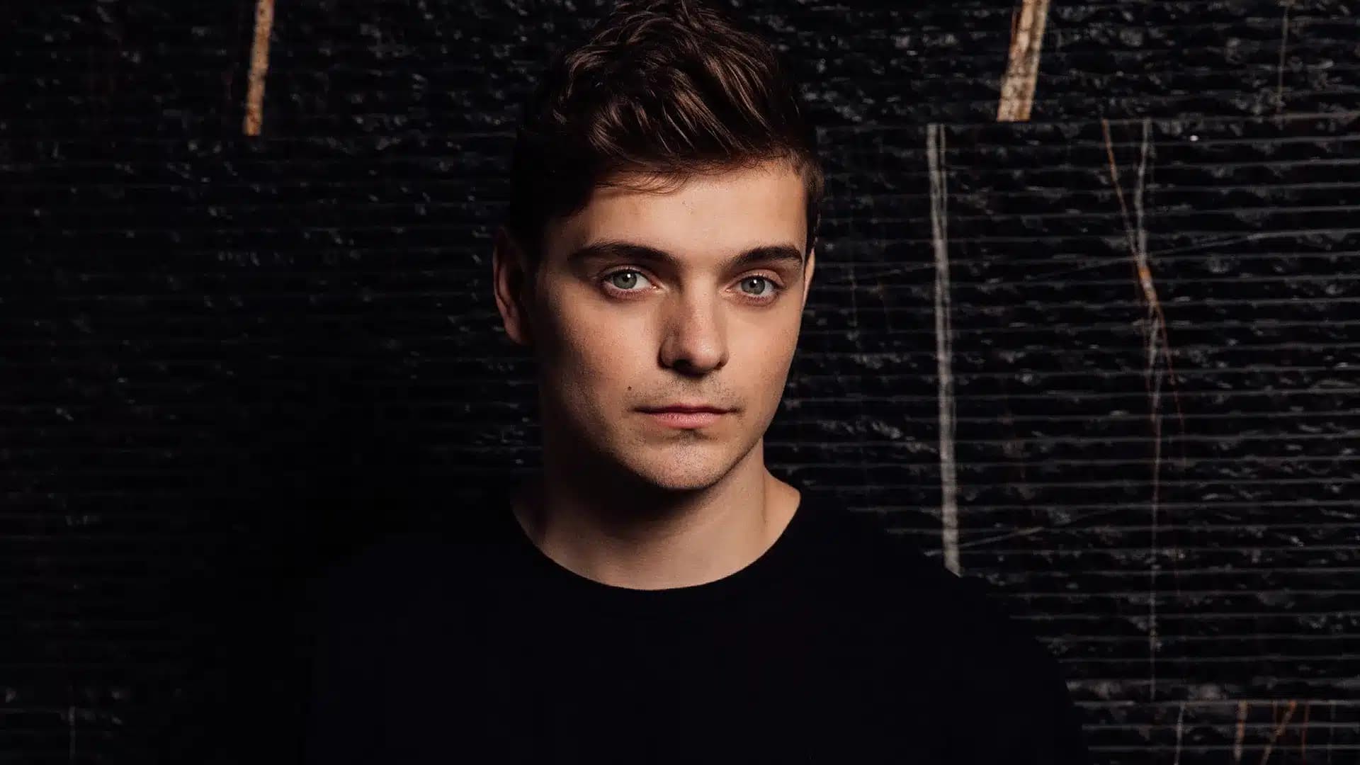 A young man with short brown hair and a serious expression, wearing a black shirt, stands against a dark, textured background.