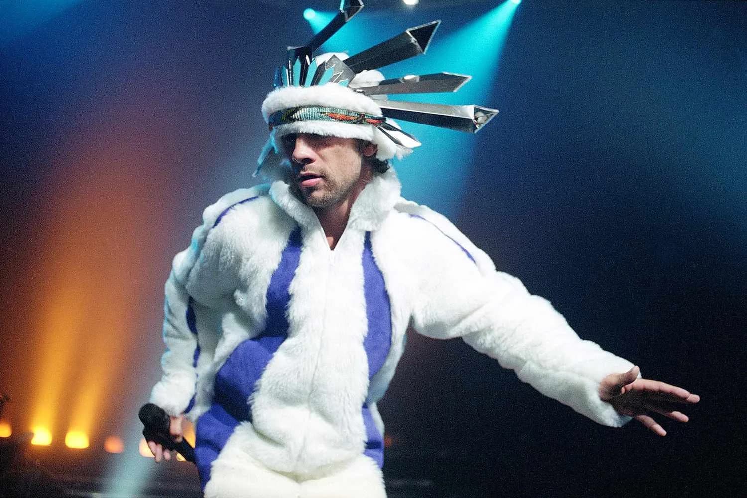 Person wearing a unique outfit with a white fur coat and a headpiece with metallic spikes, performing on stage under colorful lights.