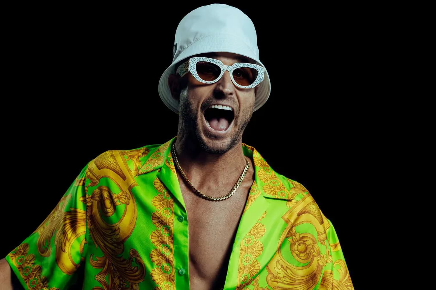 Person wearing a bright green and yellow patterned shirt, white bucket hat, and decorative sunglasses, shouting against a black background.