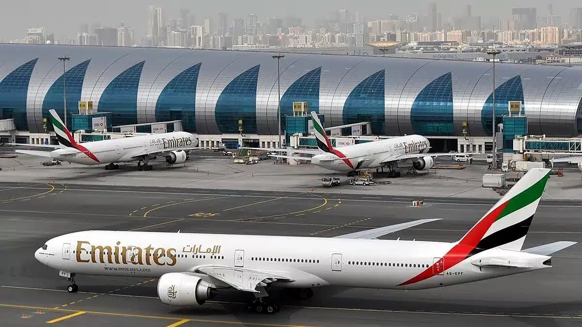 Three Emirates airplanes are parked at a busy airport terminal with a modern, arched design in the background, set against a city skyline.