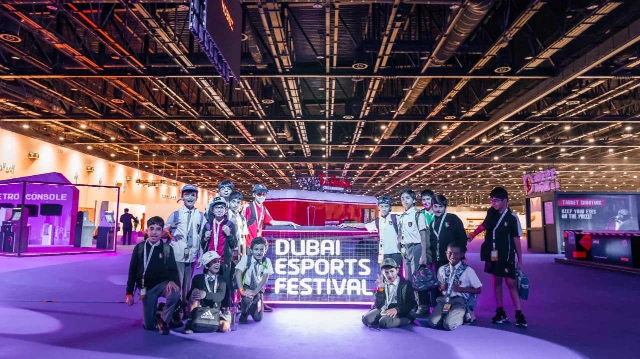 A group of children pose in front of a sign reading 'Dubai Esports Festival' inside a large, well-lit indoor venue. They appear to be excited and are dressed in casual and sporty attire, surrounded by displays and booths related to esports.