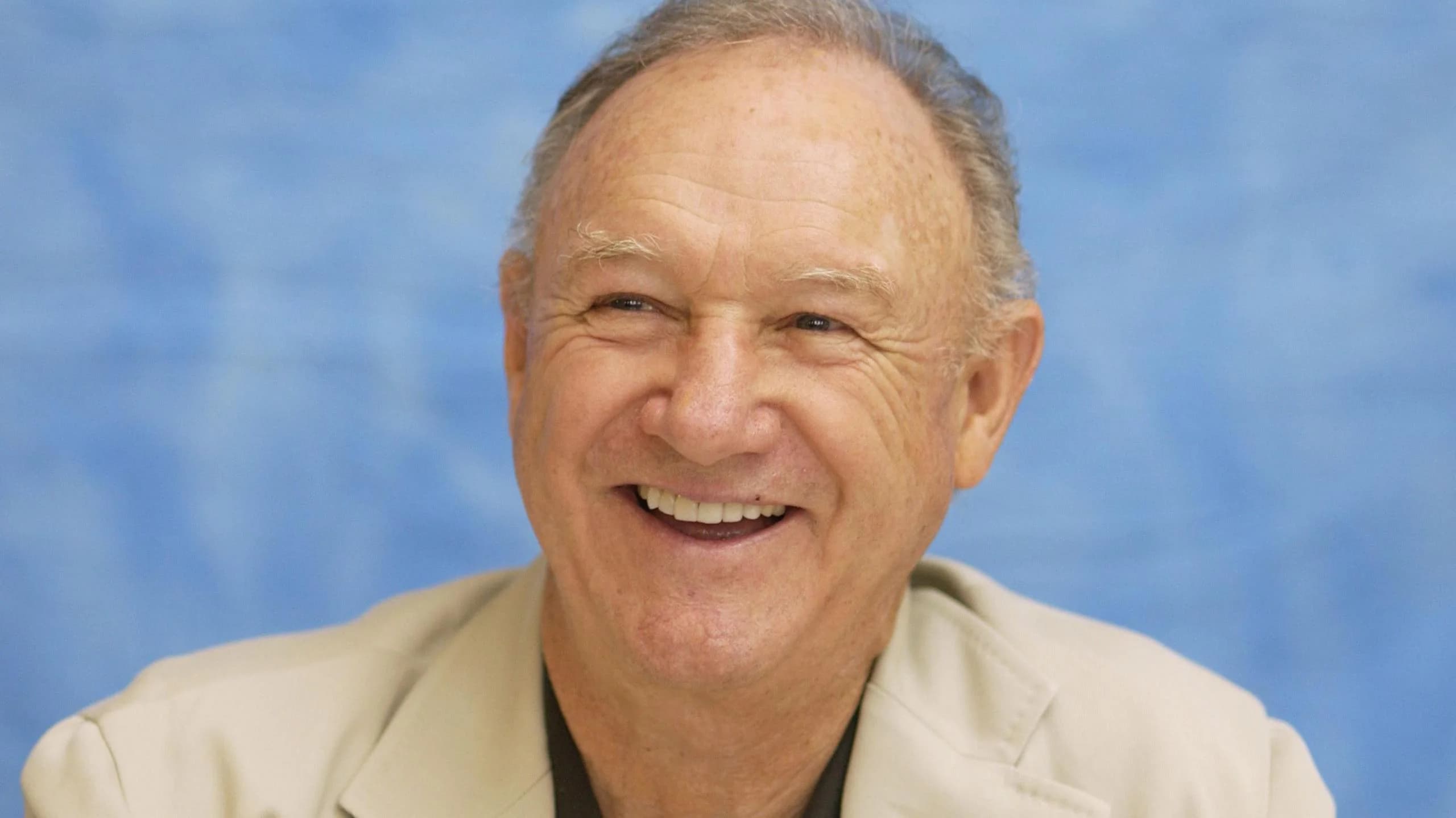 A smiling elderly man wearing a light-colored suit with a blue background.