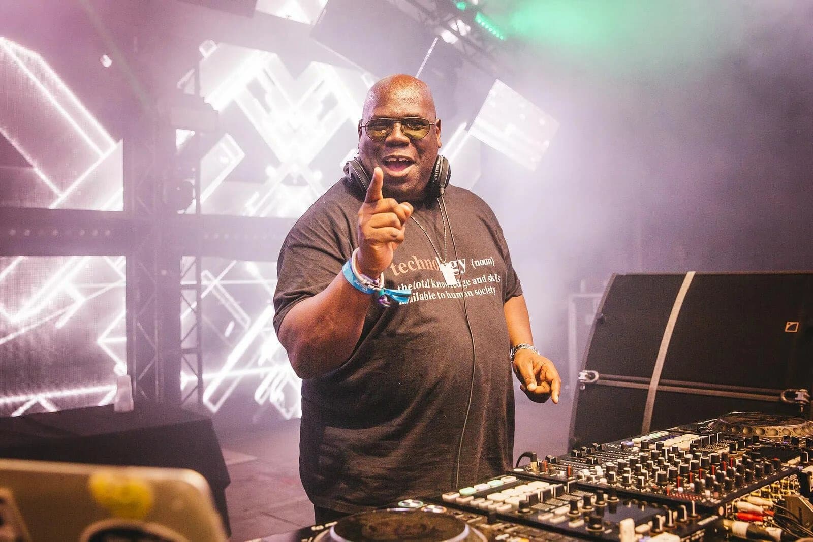 A DJ, wearing sunglasses and a black t-shirt with a definition of 'technology' printed on it, raises a finger while performing at a festival. He stands behind a DJ booth with various equipment and colorful stage lighting in a smoky atmosphere.
