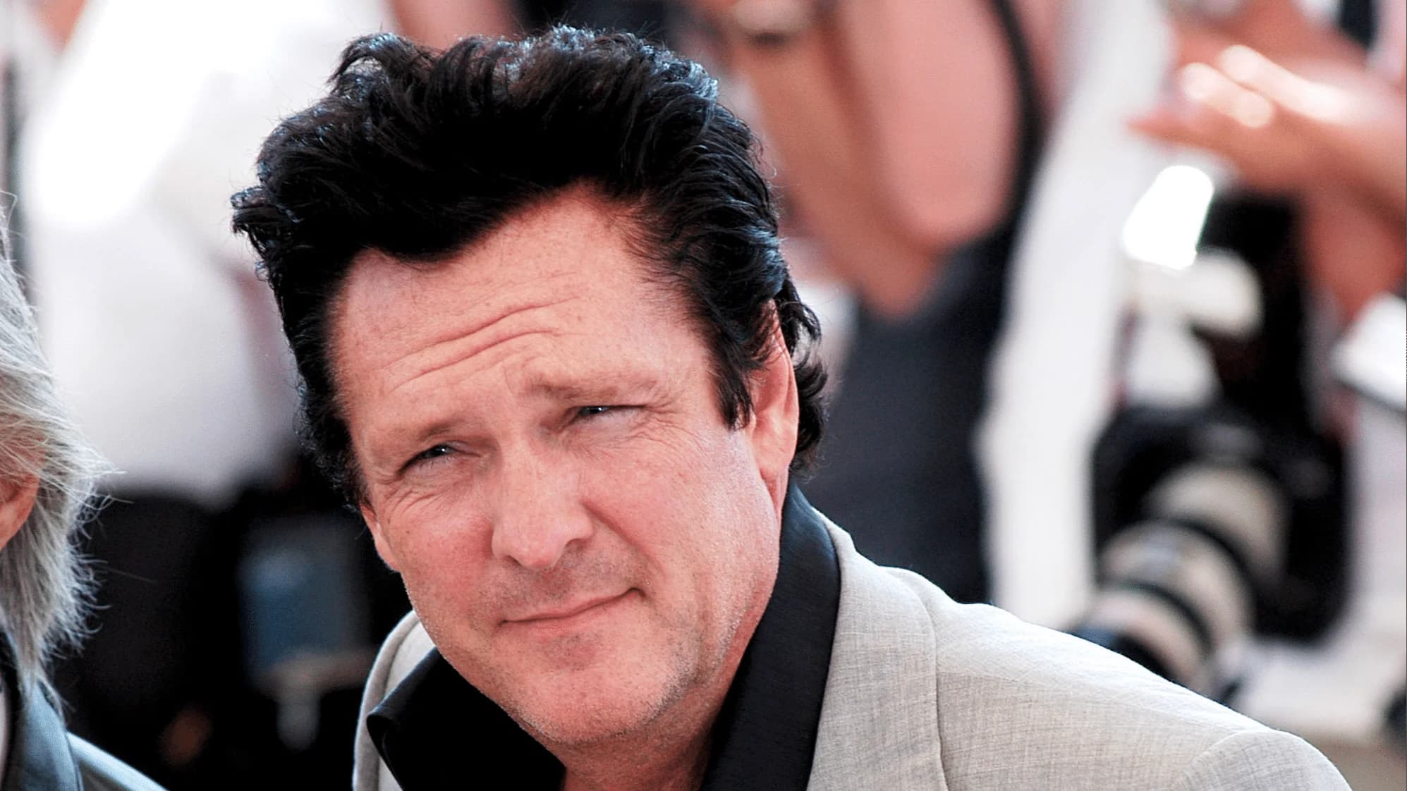 A man with short, dark, styled hair and a slight stubble is looking directly at the camera, wearing a light grey blazer over a black shirt, with soft-focus figures and cameras in the background.