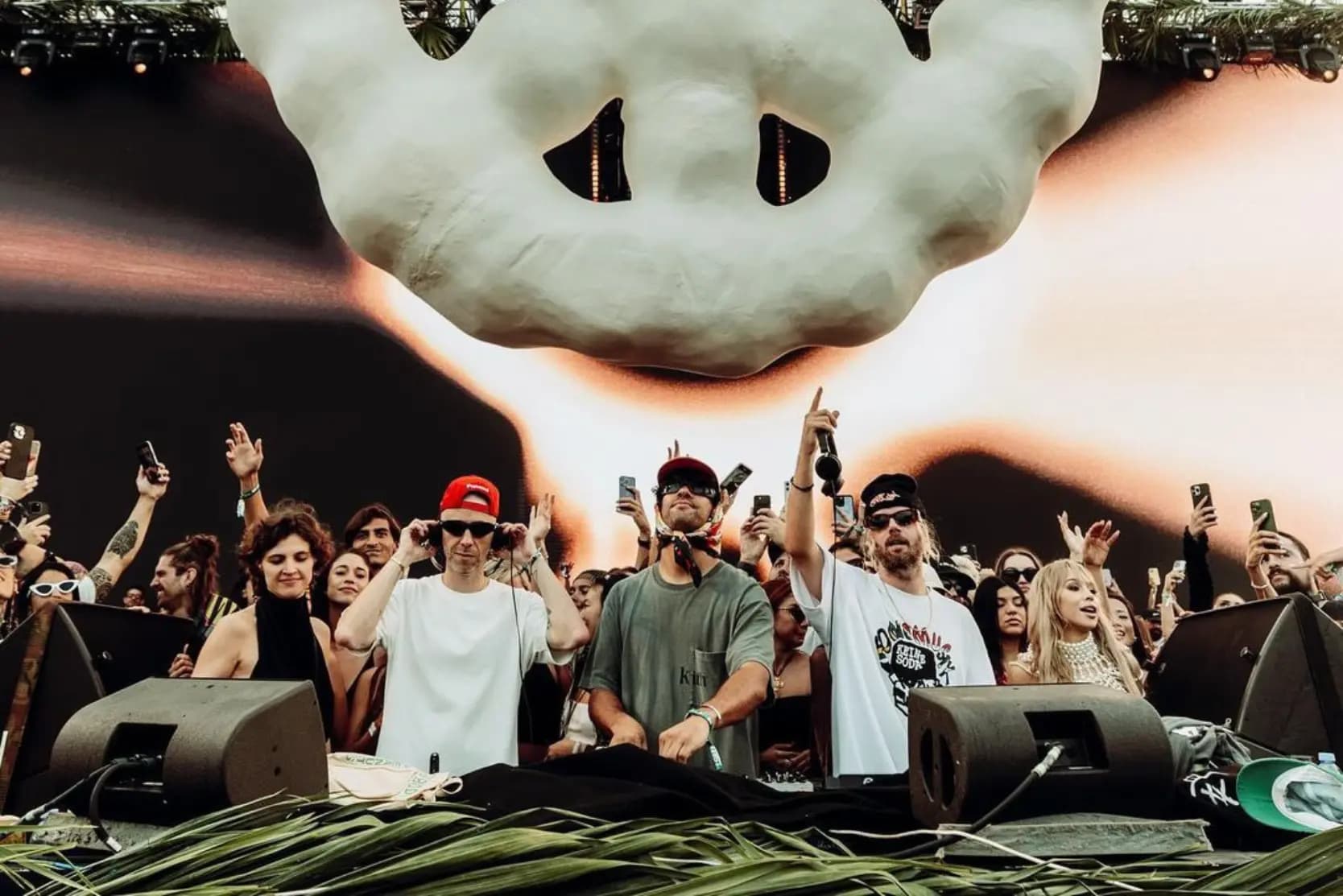 A group of people at a music event with one person in a white shirt and red cap using headphones at a DJ booth, surrounded by a crowd holding smartphones. Above them is a large, abstract cloud-like decoration.