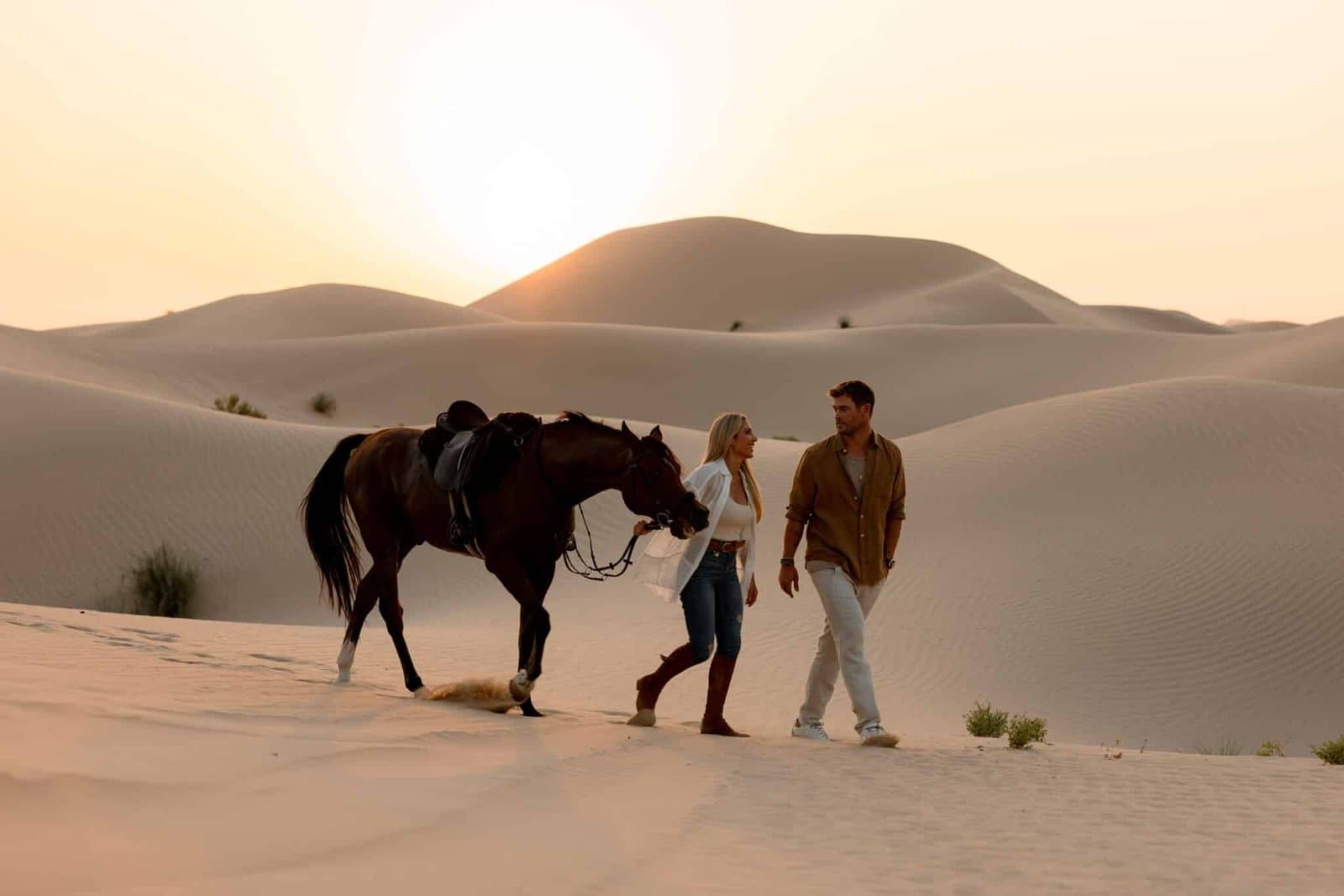 A man and woman walk alongside a horse in a desert with sand dunes at sunset.