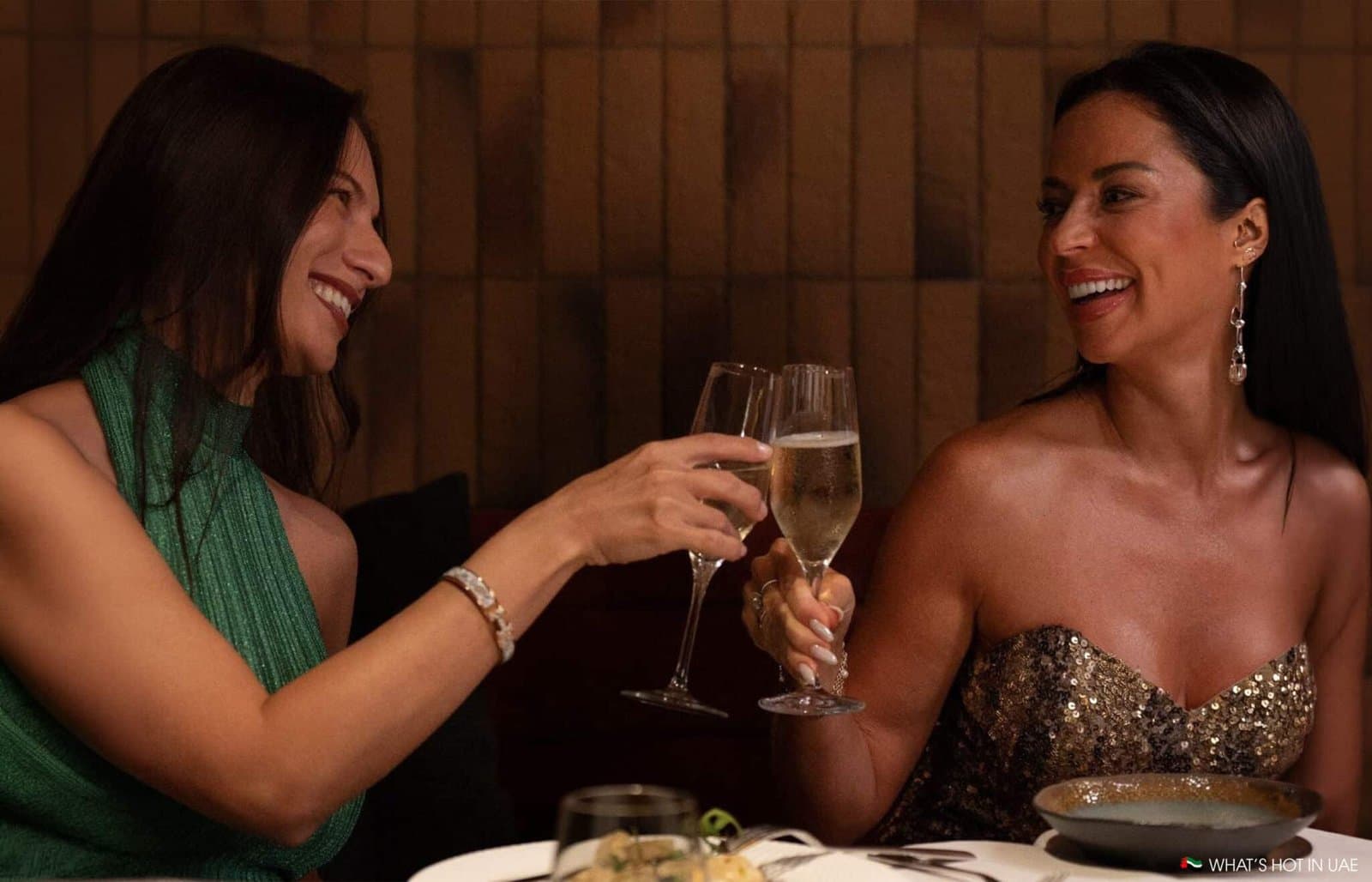 Two women smiling and clinking champagne glasses at a dining table.