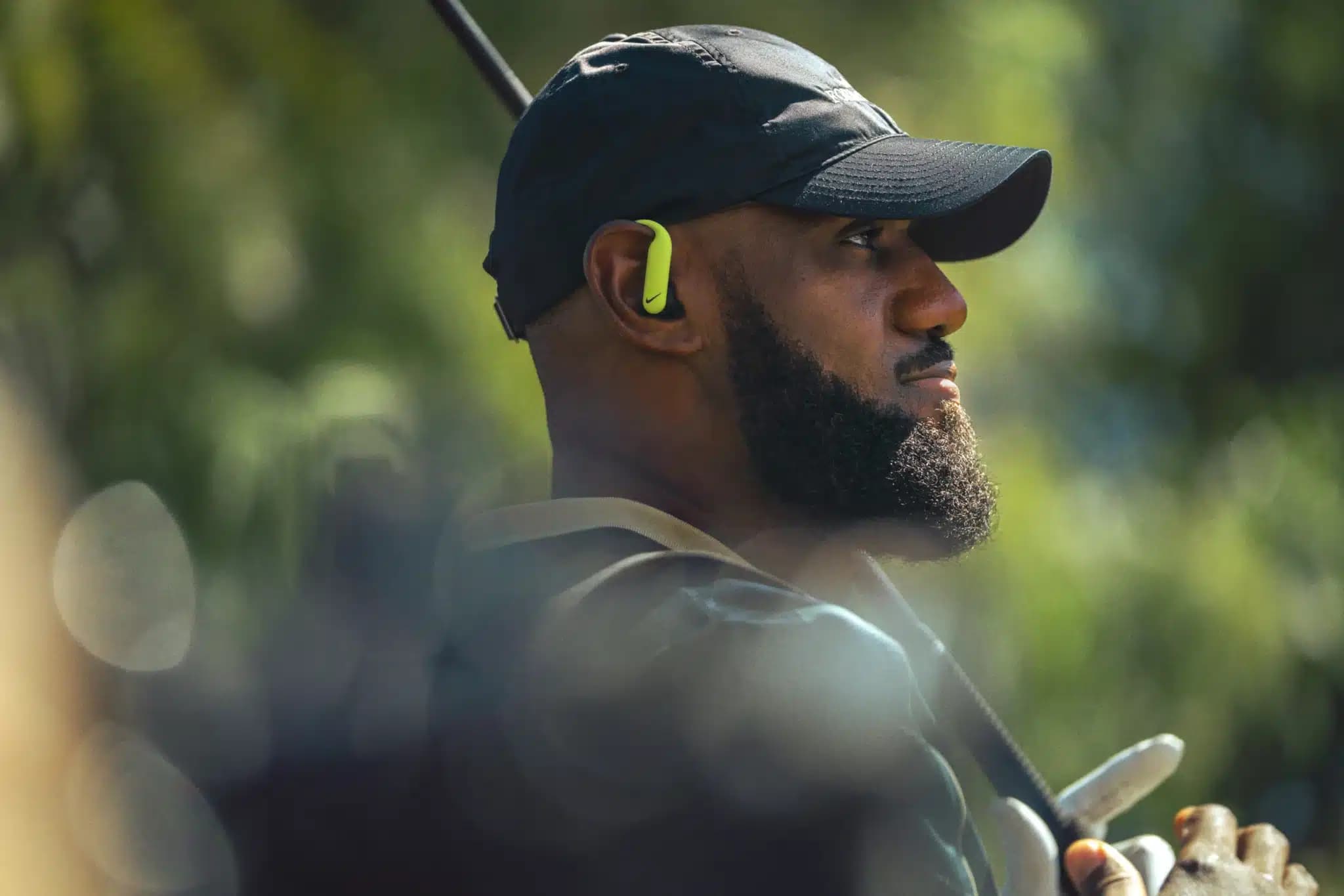 A person wearing a black cap and an earpiece, holding a golf club against a blurred outdoor background.