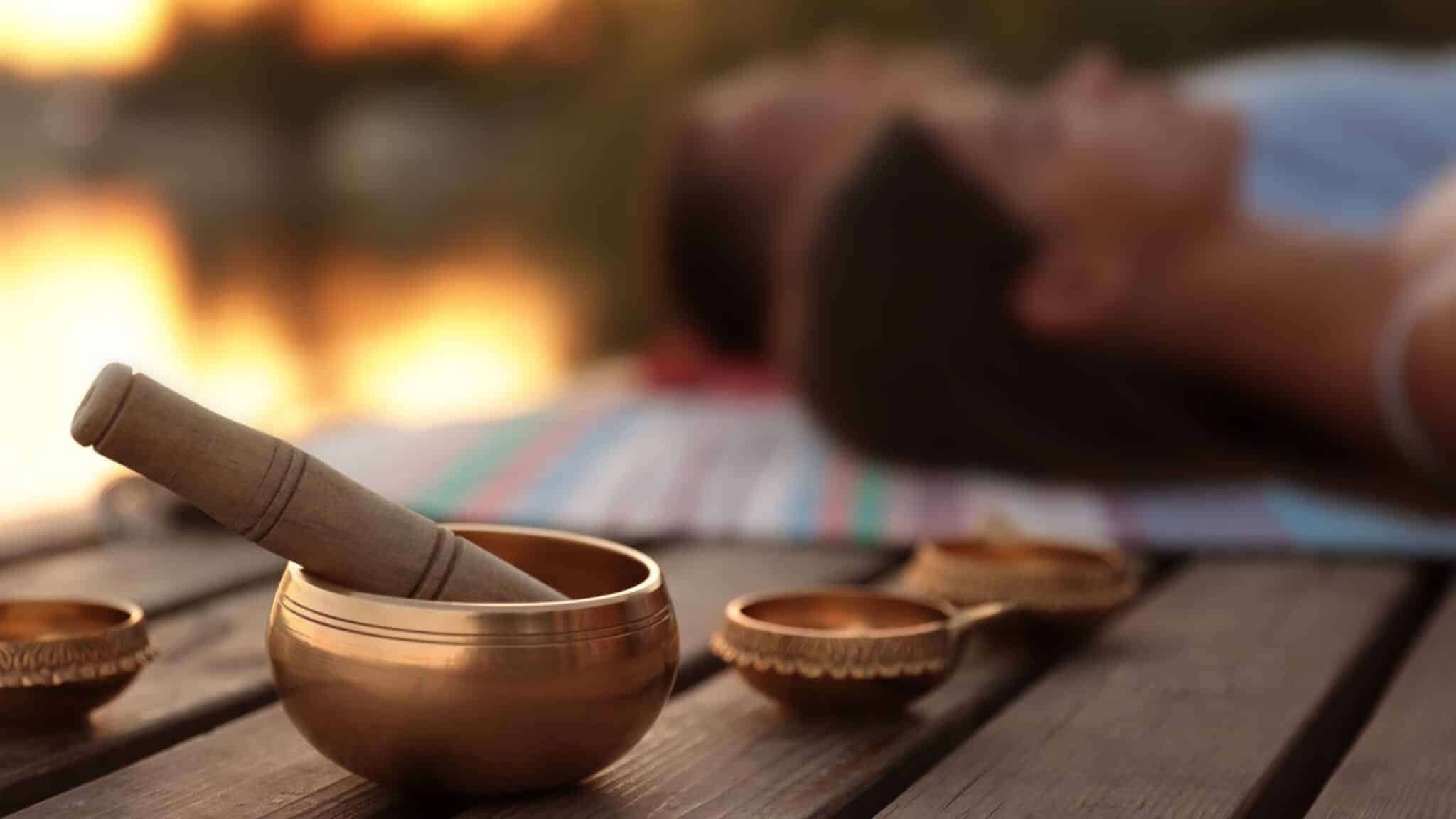 A close-up of a singing bowl with a mallet on a wooden table, with two blurred people lying on a striped blanket in the background.