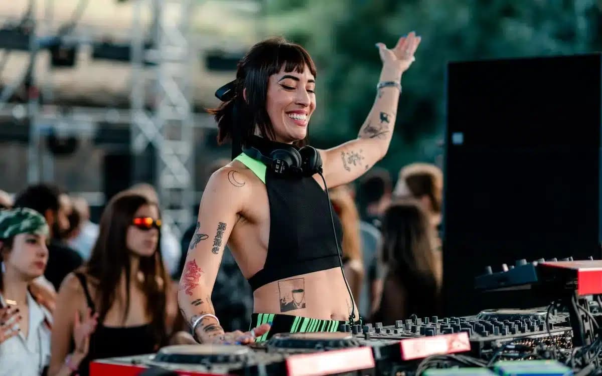 A DJ with headphones around their neck is energetically mixing music at an outdoor event, surrounded by a crowd.