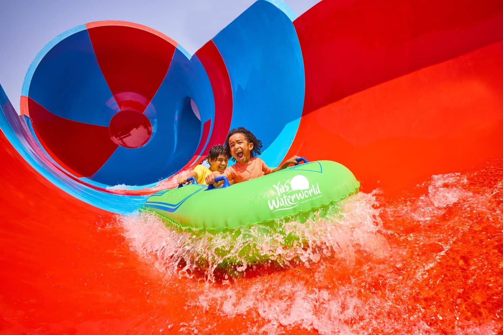 Two children excitedly ride an inflatable raft down a bright, colorful water slide.