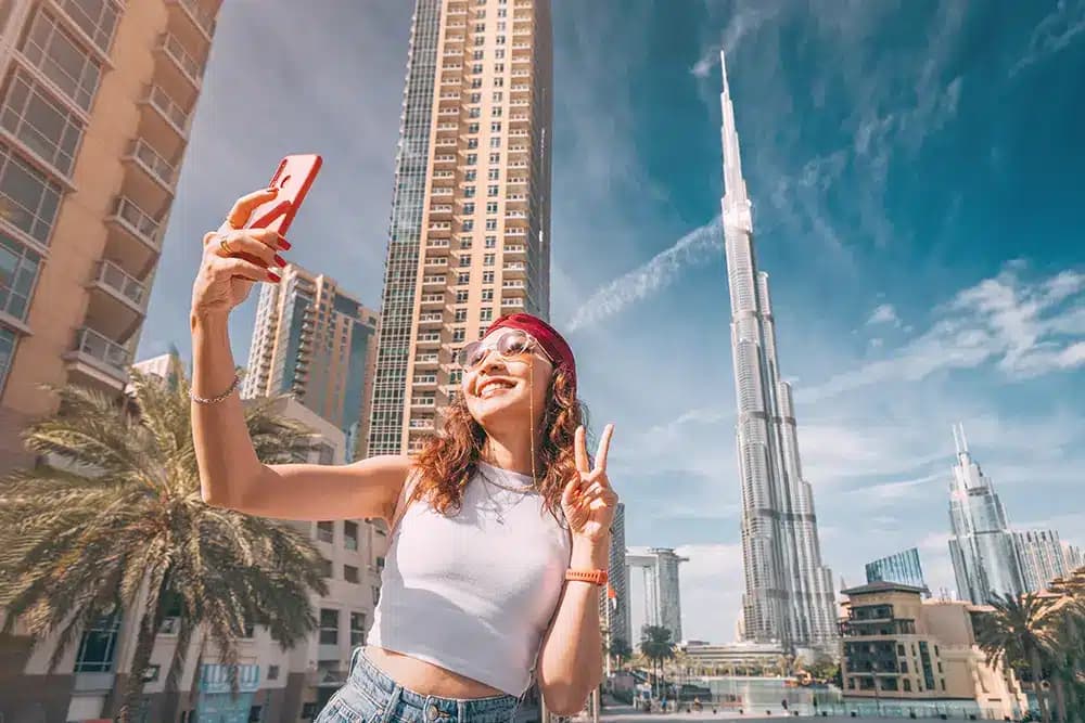 A woman takes a selfie near tall skyscrapers, including the Burj Khalifa, on a sunny day.