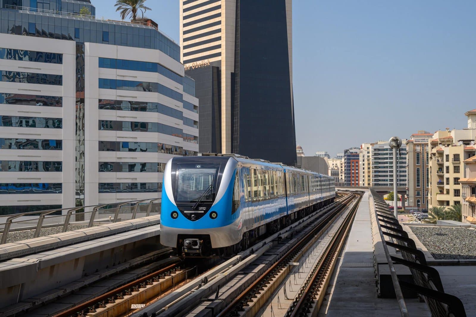 A blue and silver metro train travels on an elevated track between modern high-rise buildings on a clear day.