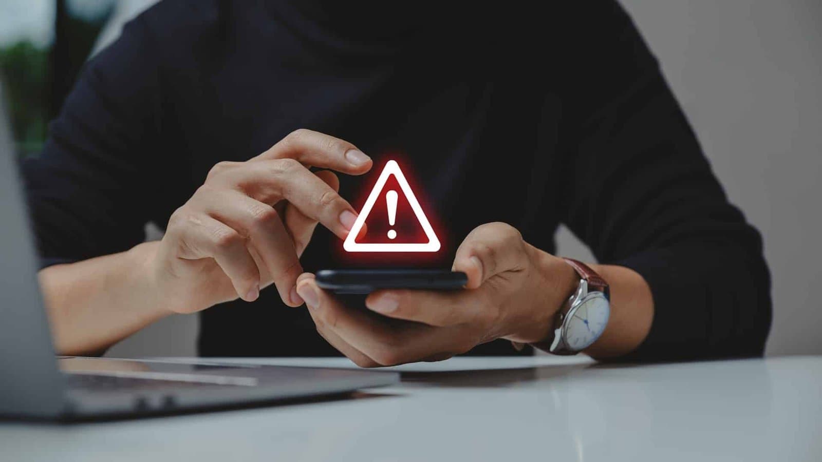 A person holding a smartphone with a glowing warning icon above it, seated at a desk with a laptop.