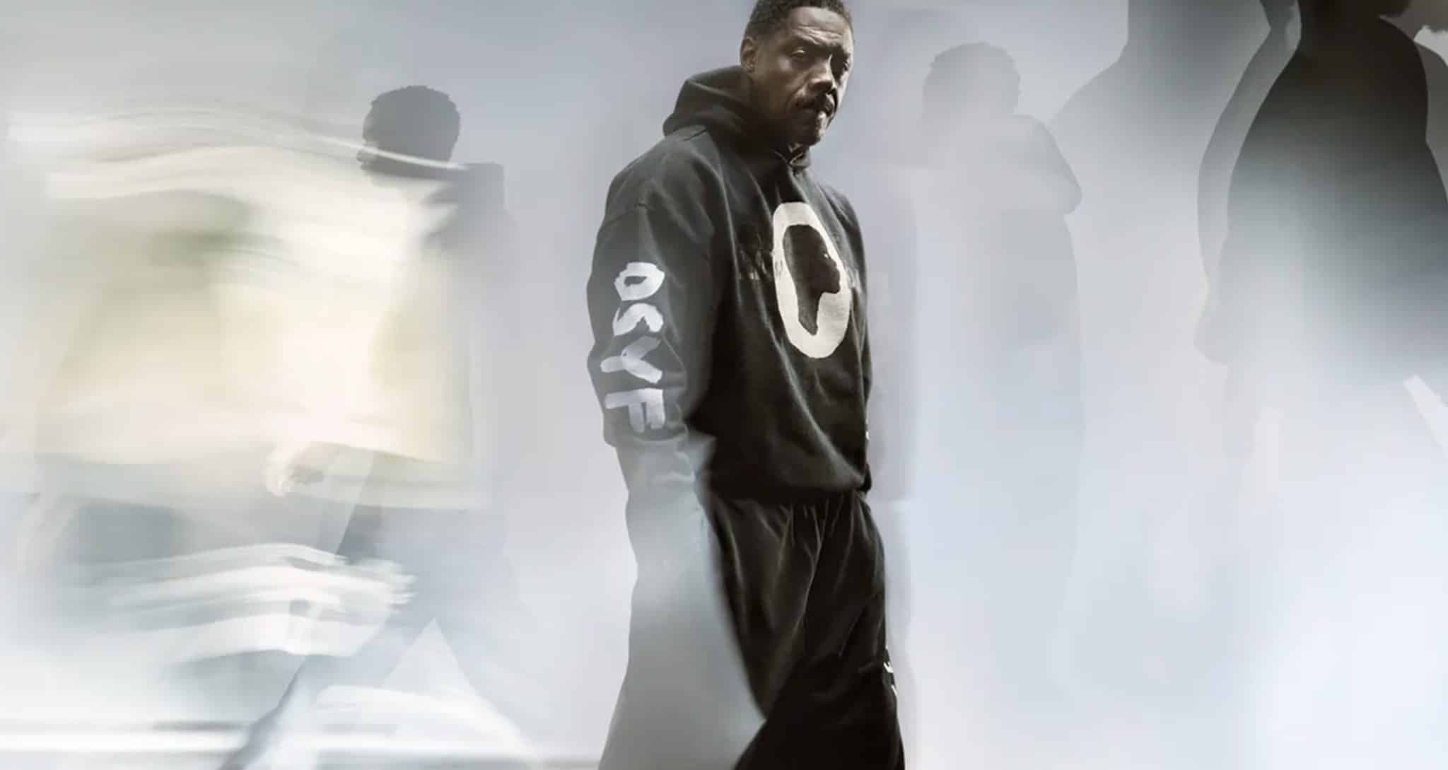 A man in a black hoodie with "OSYF" on the sleeve stands still amid blurred figures in motion.