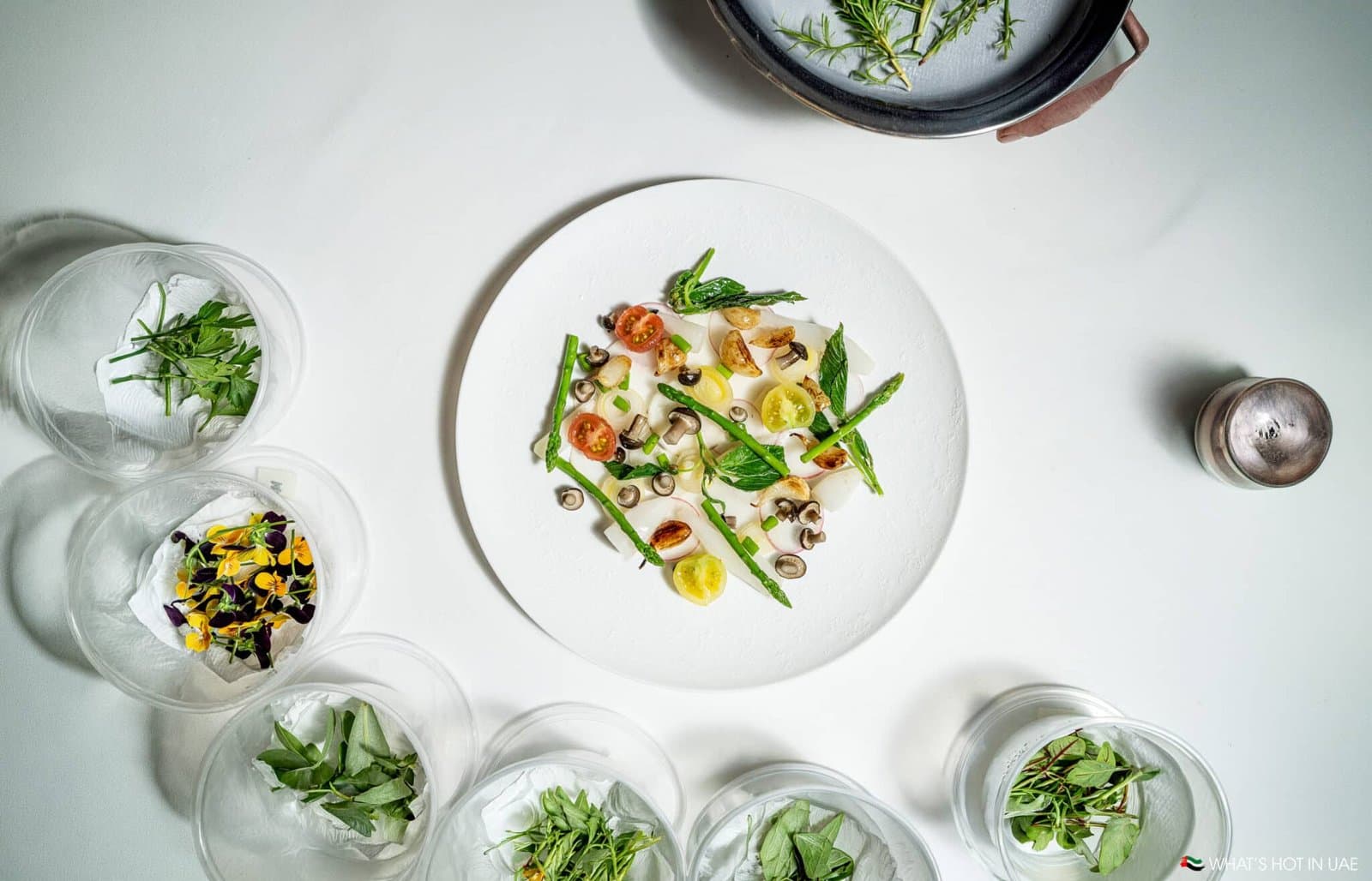 Overhead view of a plated gourmet vegetable dish surrounded by bowls of fresh herbs and edible flowers