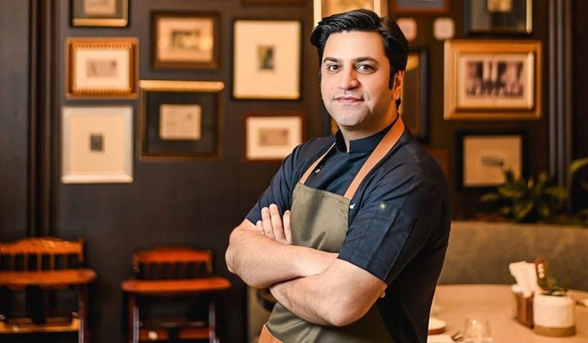 A person in a chef's apron stands confidently with arms crossed in a warmly lit room decorated with framed pictures on the wall.