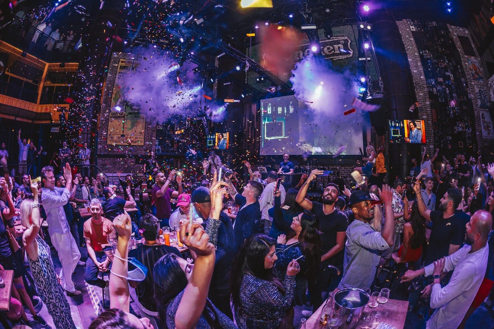 Crowded nightclub with confetti, smoke, and people cheering under colorful stage lights