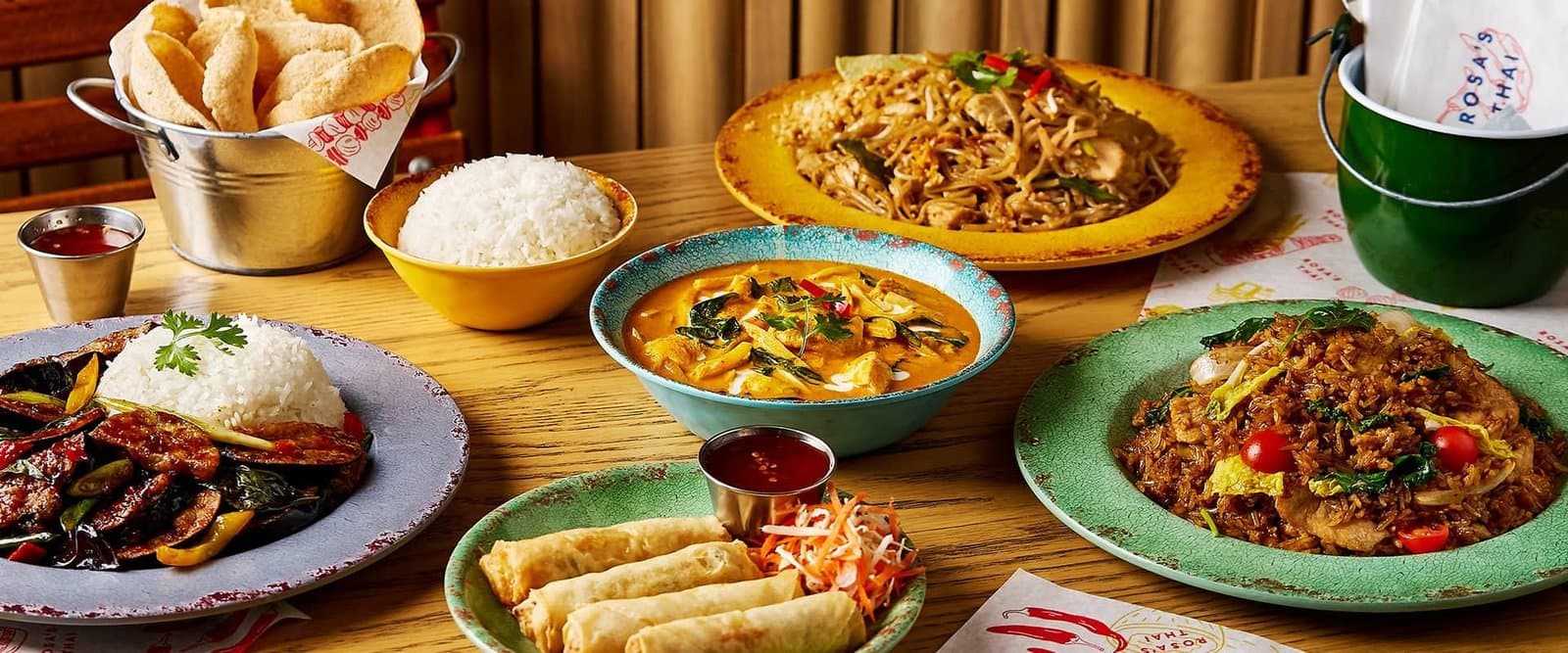 A spread of various Asian dishes including rice, curry, spring rolls, and stir-fried noodles on a wooden table.