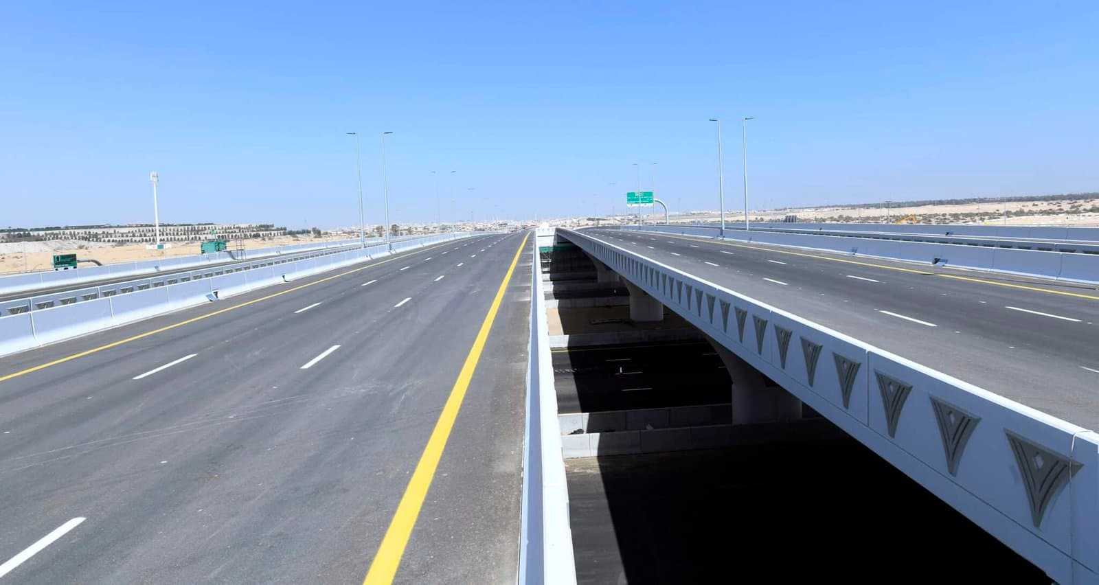 A wide, empty highway with two parallel bridges under a clear blue sky.