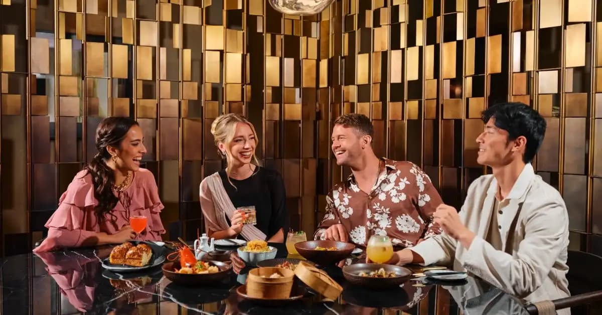 Four friends laugh over drinks and dishes at a restaurant table with a gold geometric wall behind them