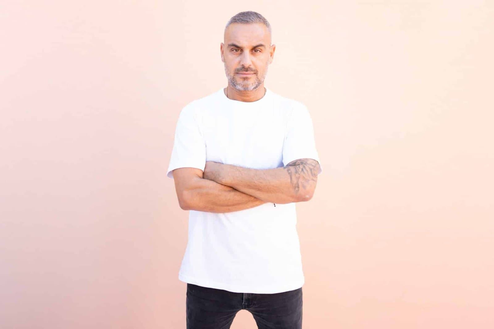 Man with short gray hair and a tattooed arm, wearing a white T-shirt and black jeans, standing with arms crossed against a pink wall