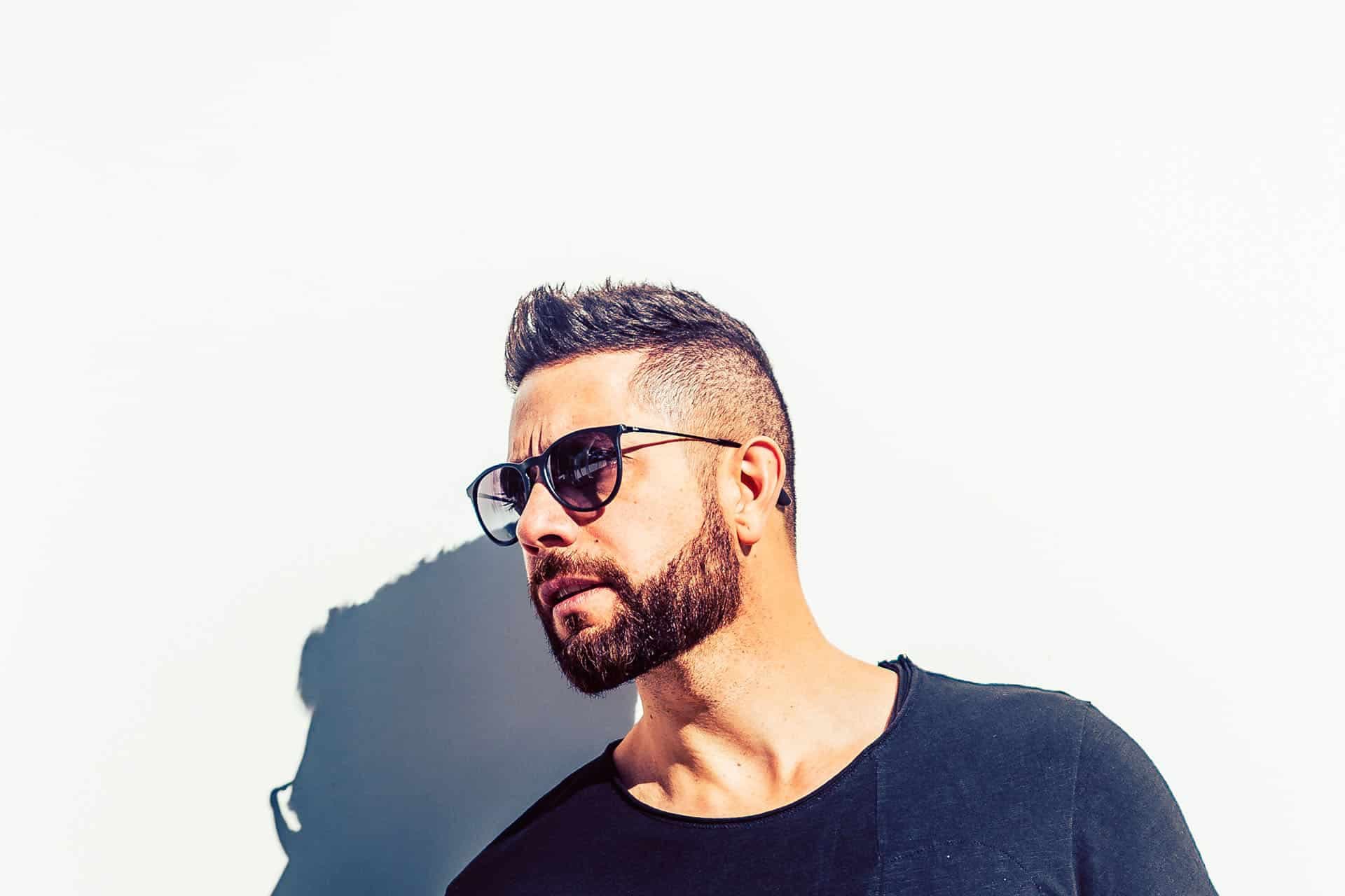 Man with beard and sunglasses in a black shirt against a white wall, with a strong shadow behind him