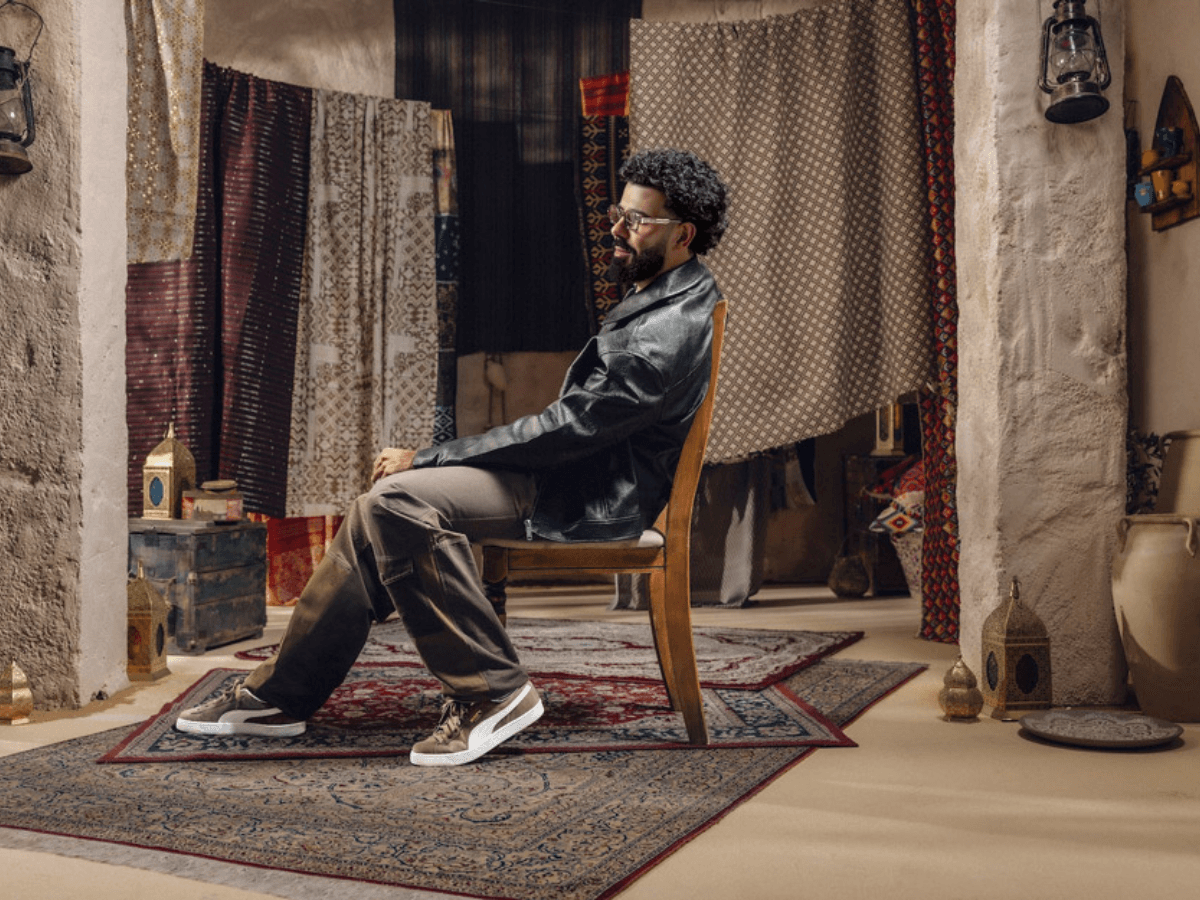 A man in a black leather jacket sits on a chair in a room with hanging patterned fabrics and rugs