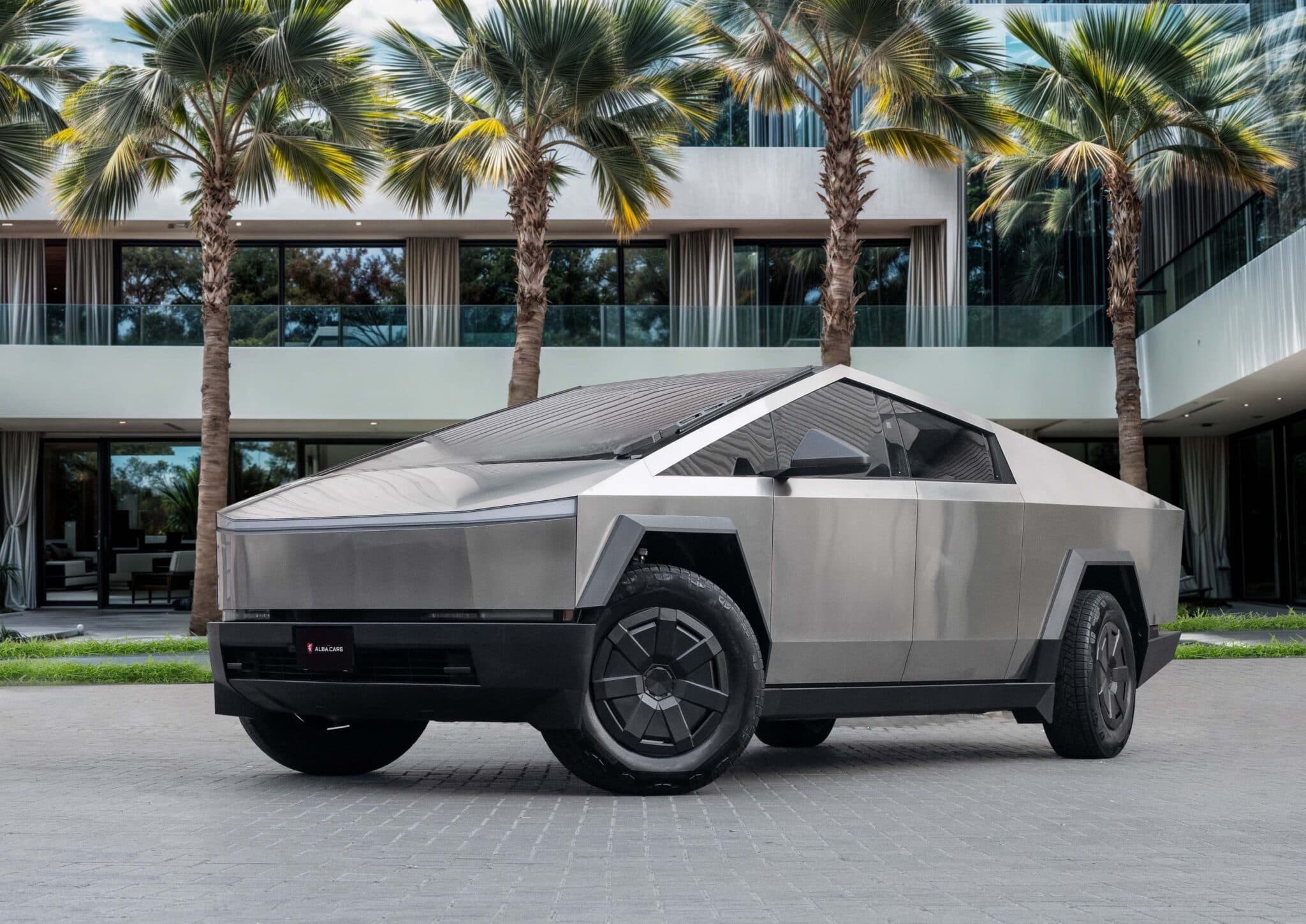 Angular silver electric pickup truck parked in front of a modern house lined with palm trees