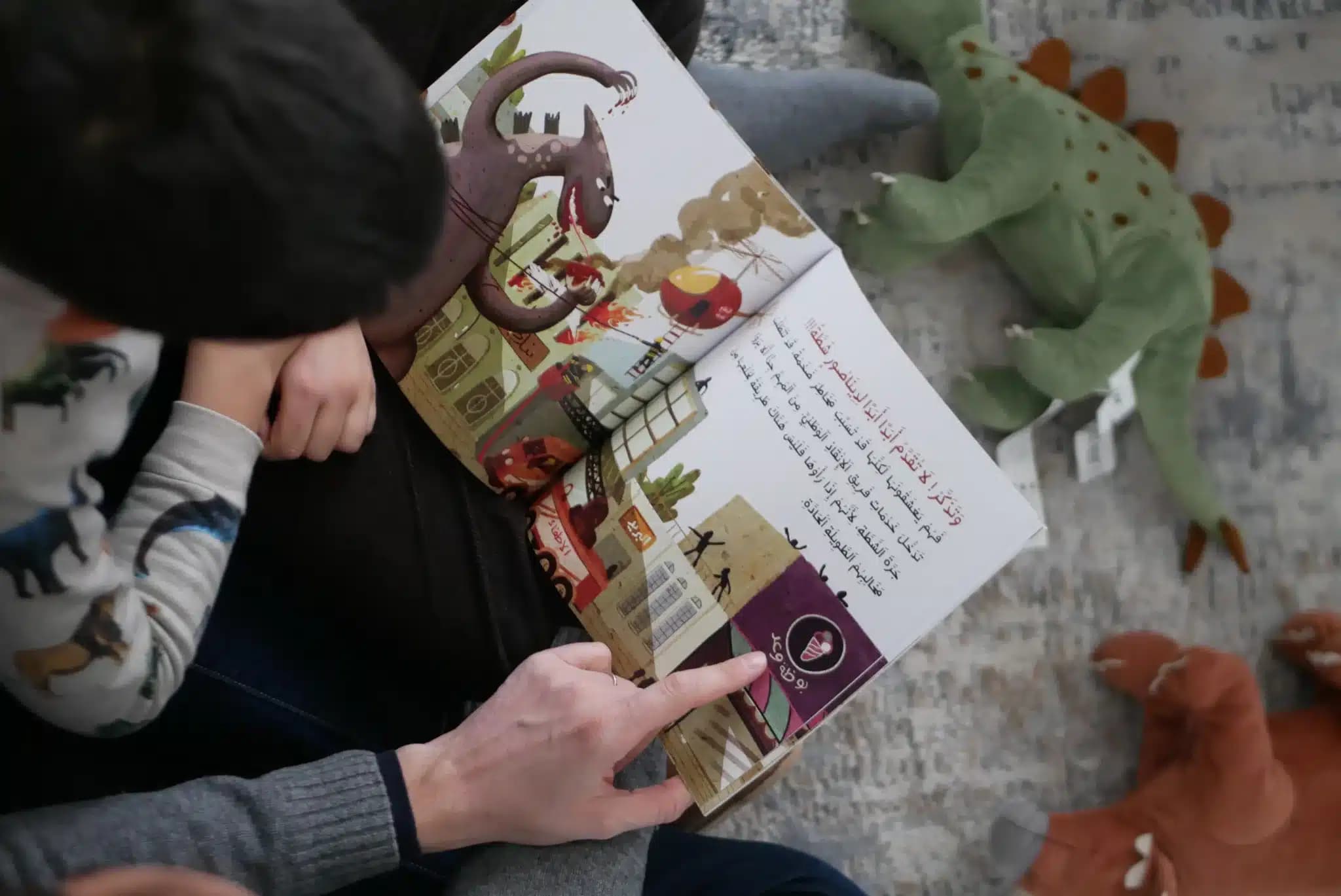 Adult reads an Arabic picture book to a child surrounded by stuffed dinosaur toys