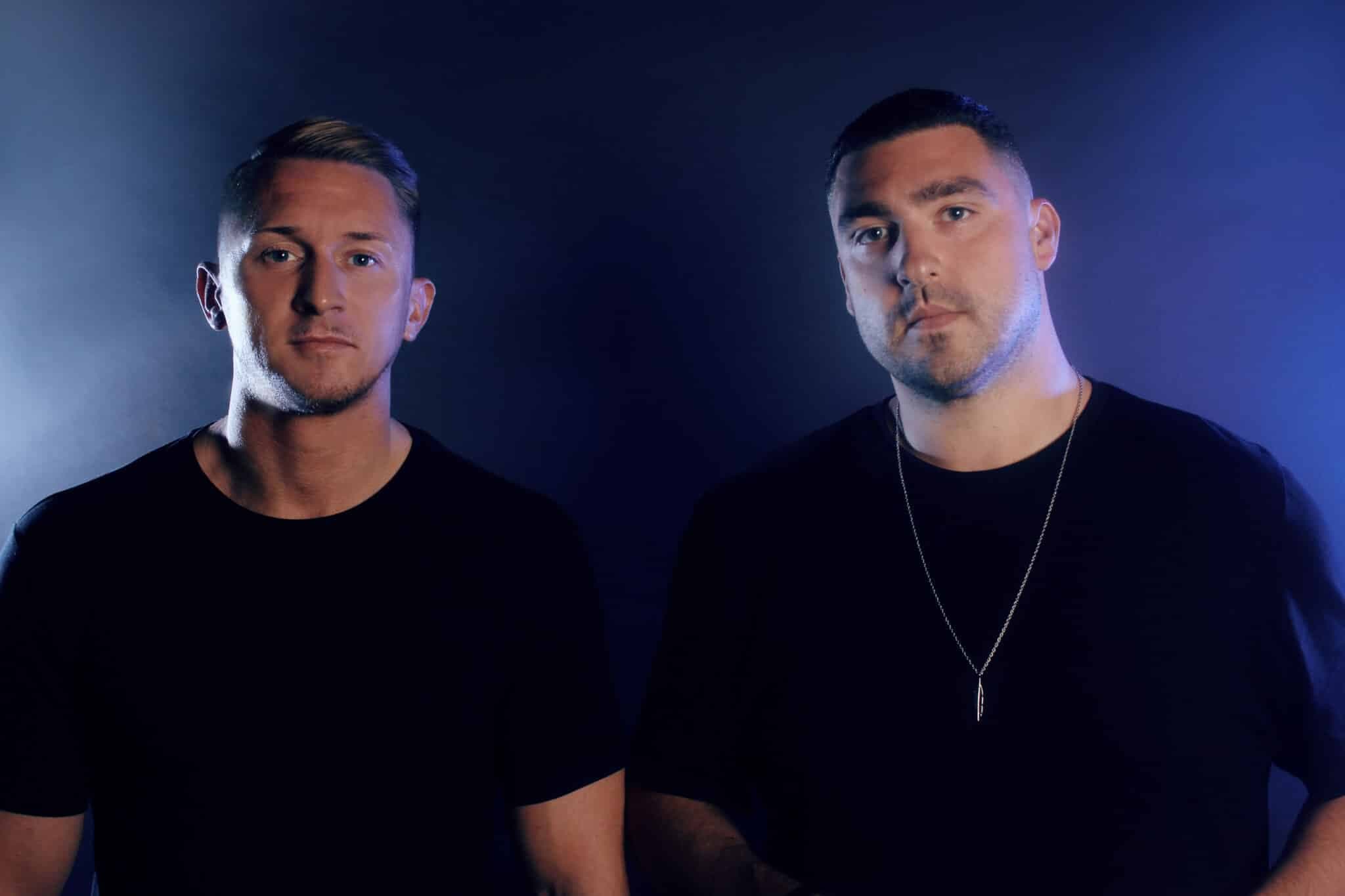 Two men in black shirts pose against a dark blue background, with the man on the right wearing a pendant necklace