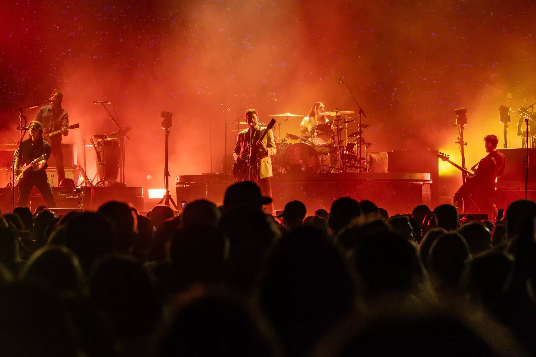 Band performing onstage under orange lighting, with a singer and guitarists before a large concert crowd