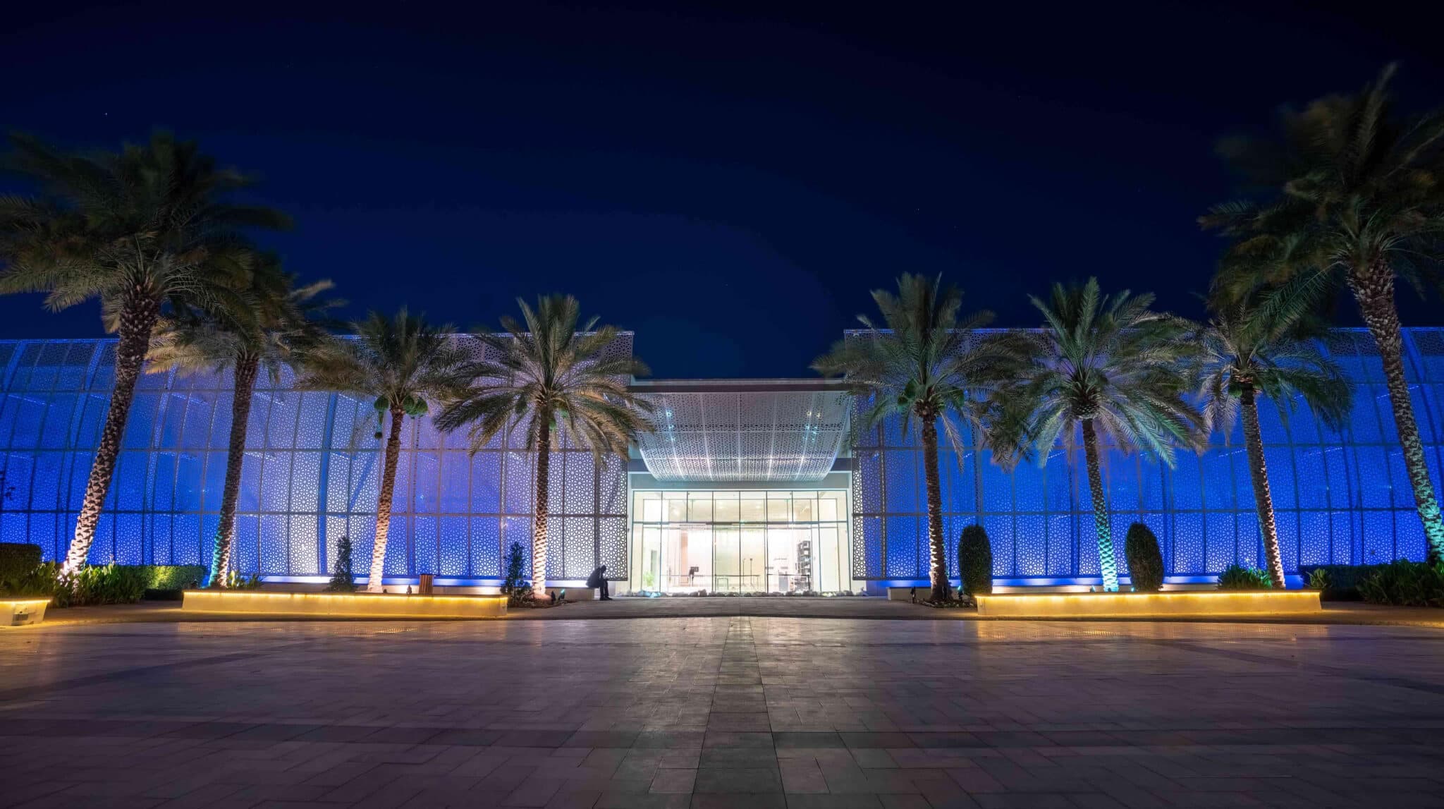 Nighttime view of a modern building entrance illuminated blue, framed by palm trees and patterned screens
