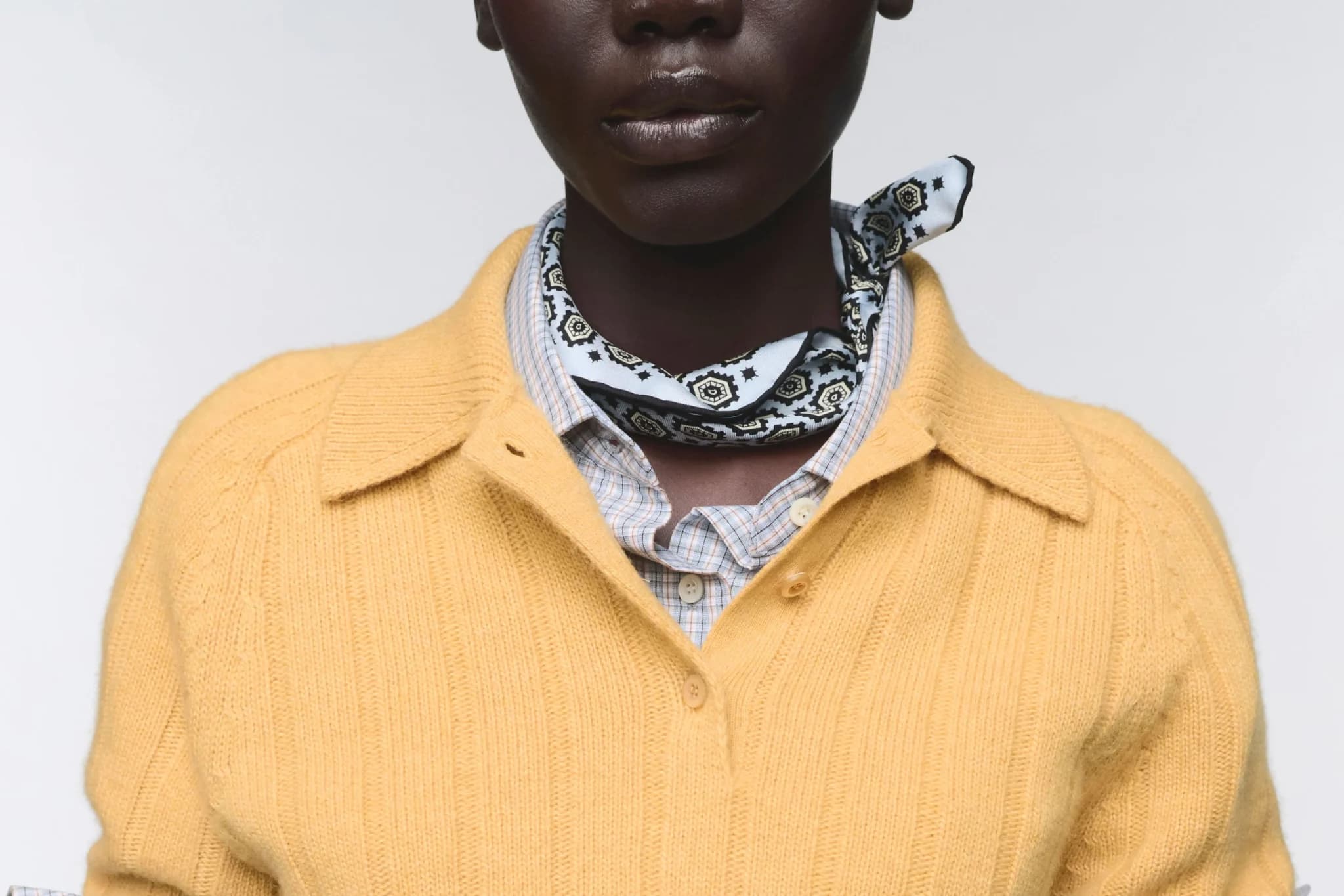 Person wearing a yellow cardigan, plaid shirt, and black-and-white patterned neckerchief