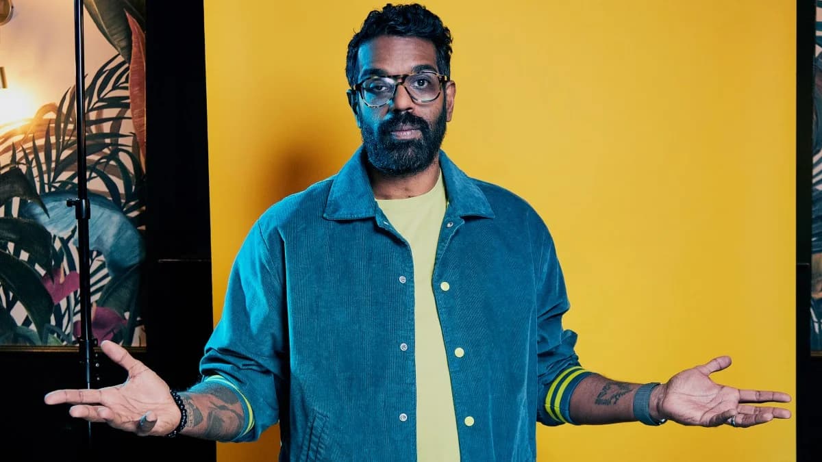 A man with glasses and a beard wearing a blue jacket and yellow shirt, standing against a bright yellow background with his arms outstretched.