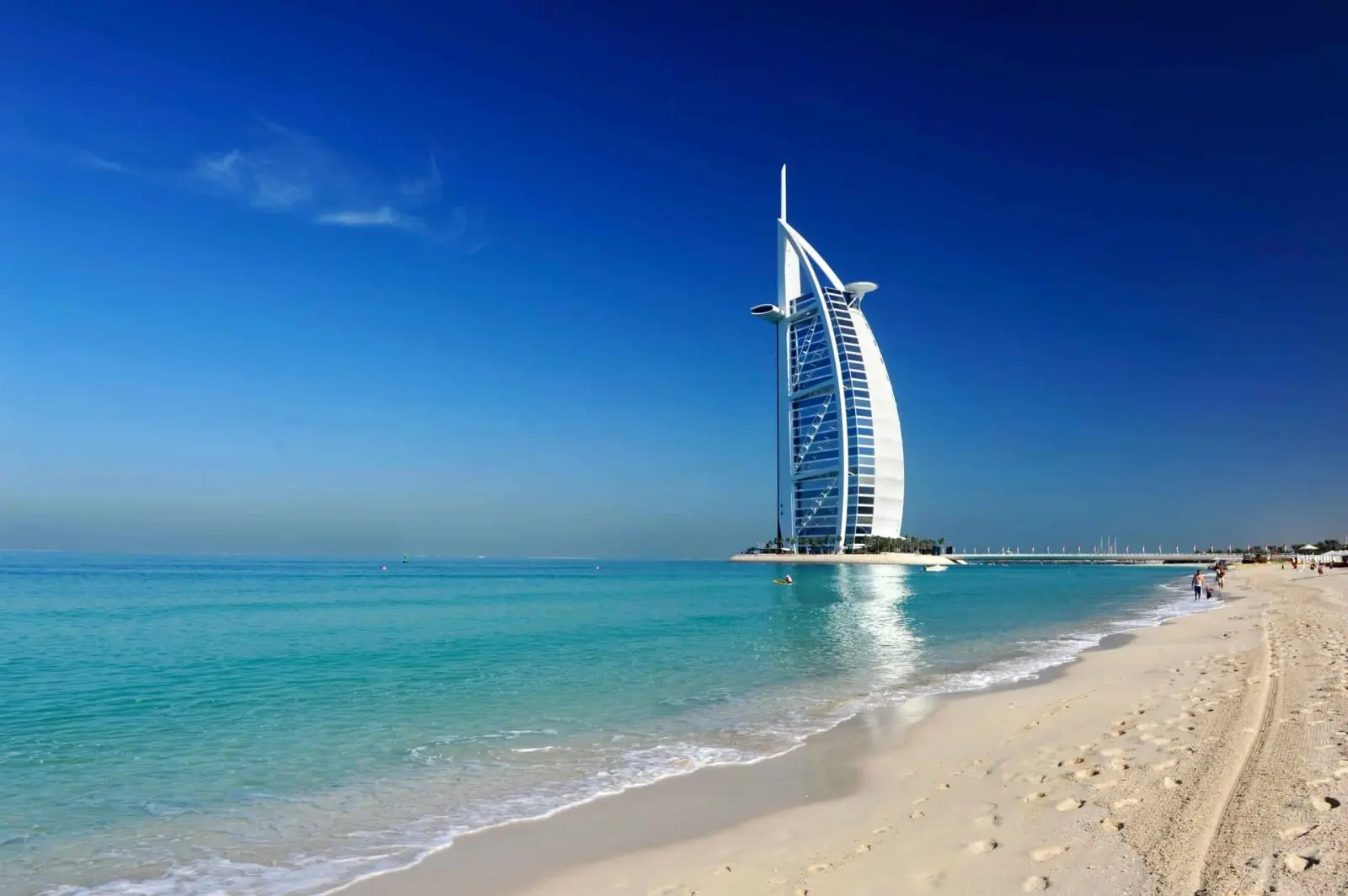 A modern sail-shaped skyscraper near a sandy beach with turquoise water under a clear blue sky.