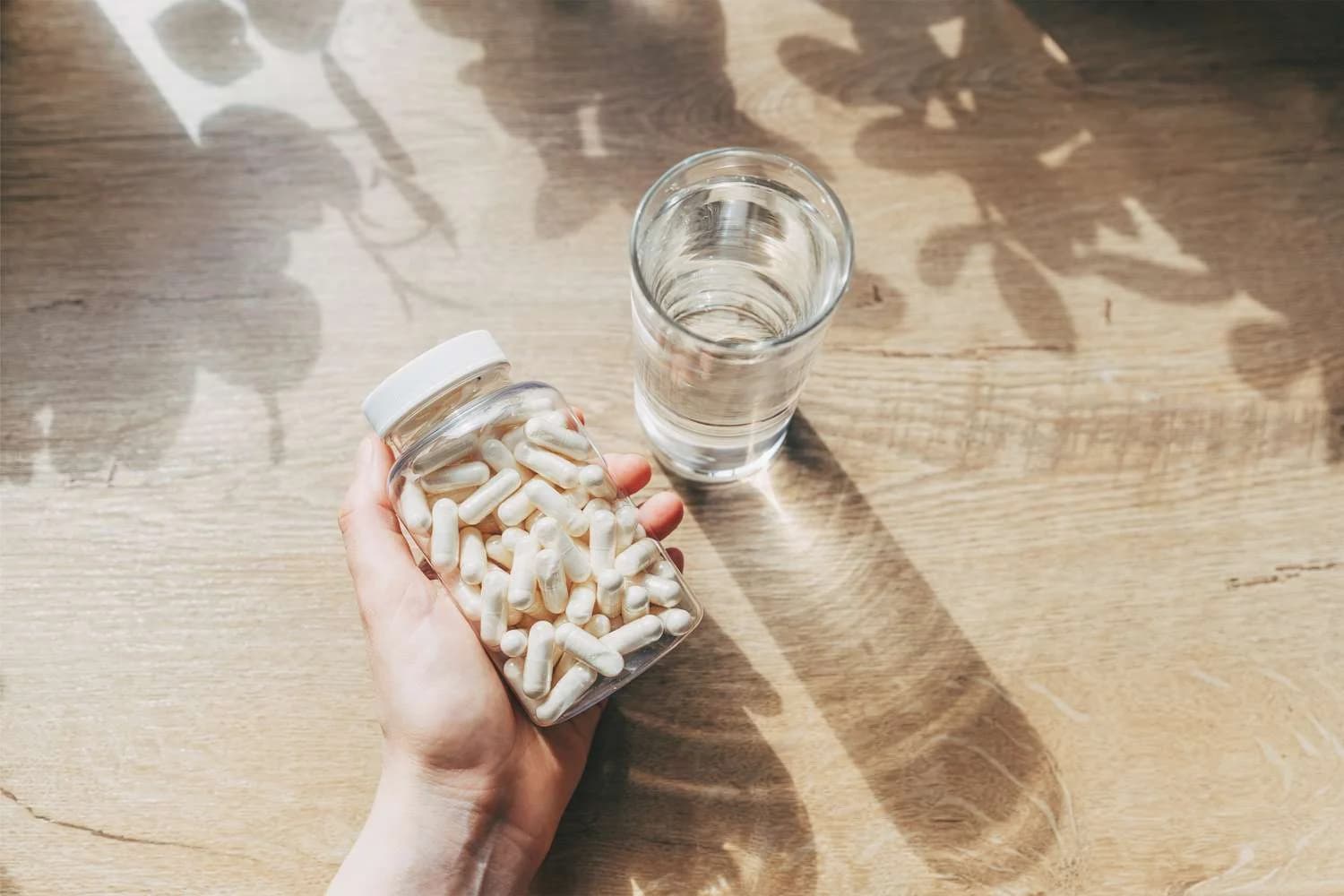 A hand holding a clear jar filled with white capsules next to a glass of water on a wooden surface, with leaf shadows cast on the table.