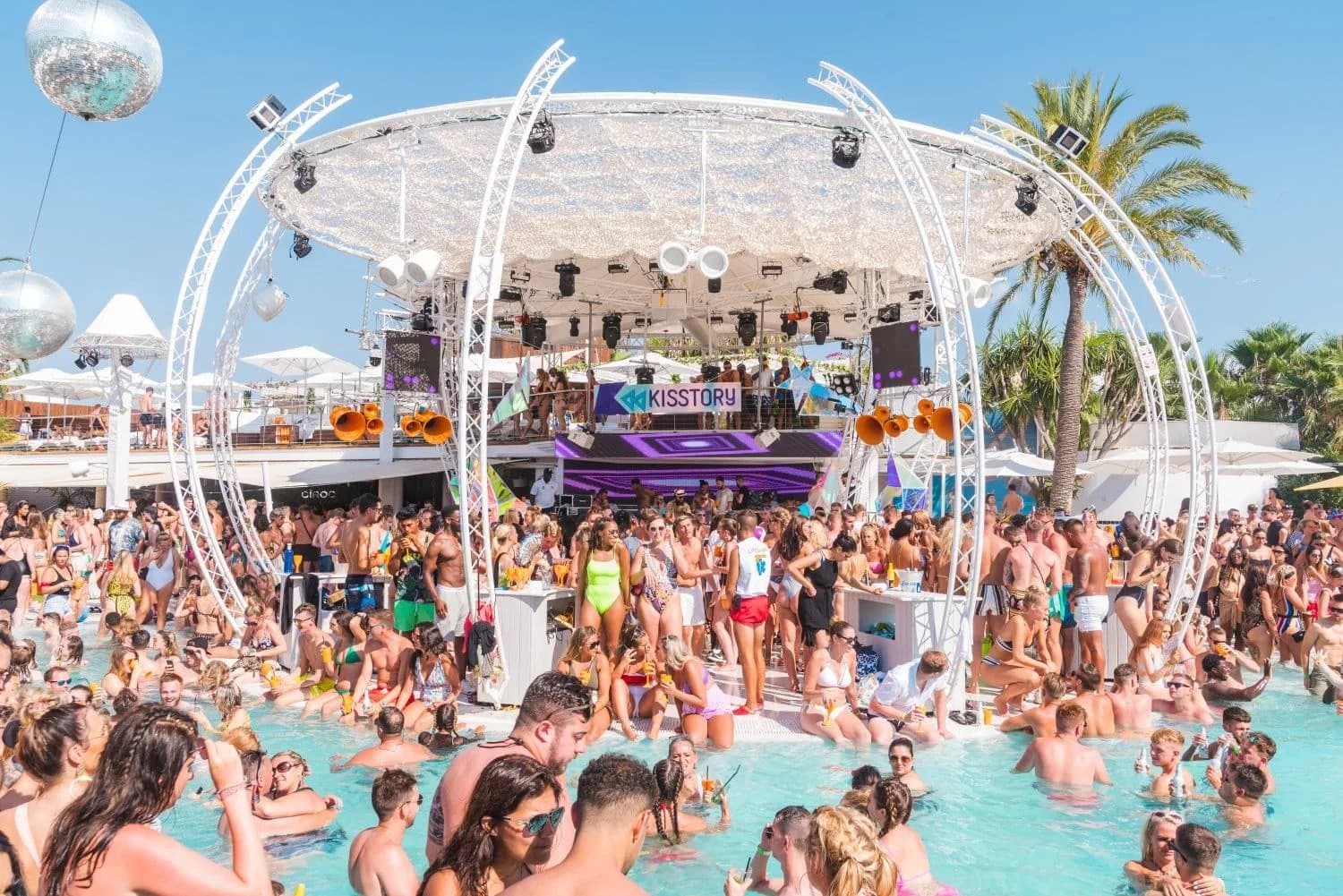 A lively pool party with a large crowd of people enjoying the sun, swimming, and dancing around a DJ stage with a "Kisstory" sign. The scene includes decorative structures, palm trees, and a vibrant atmosphere under a clear blue sky.