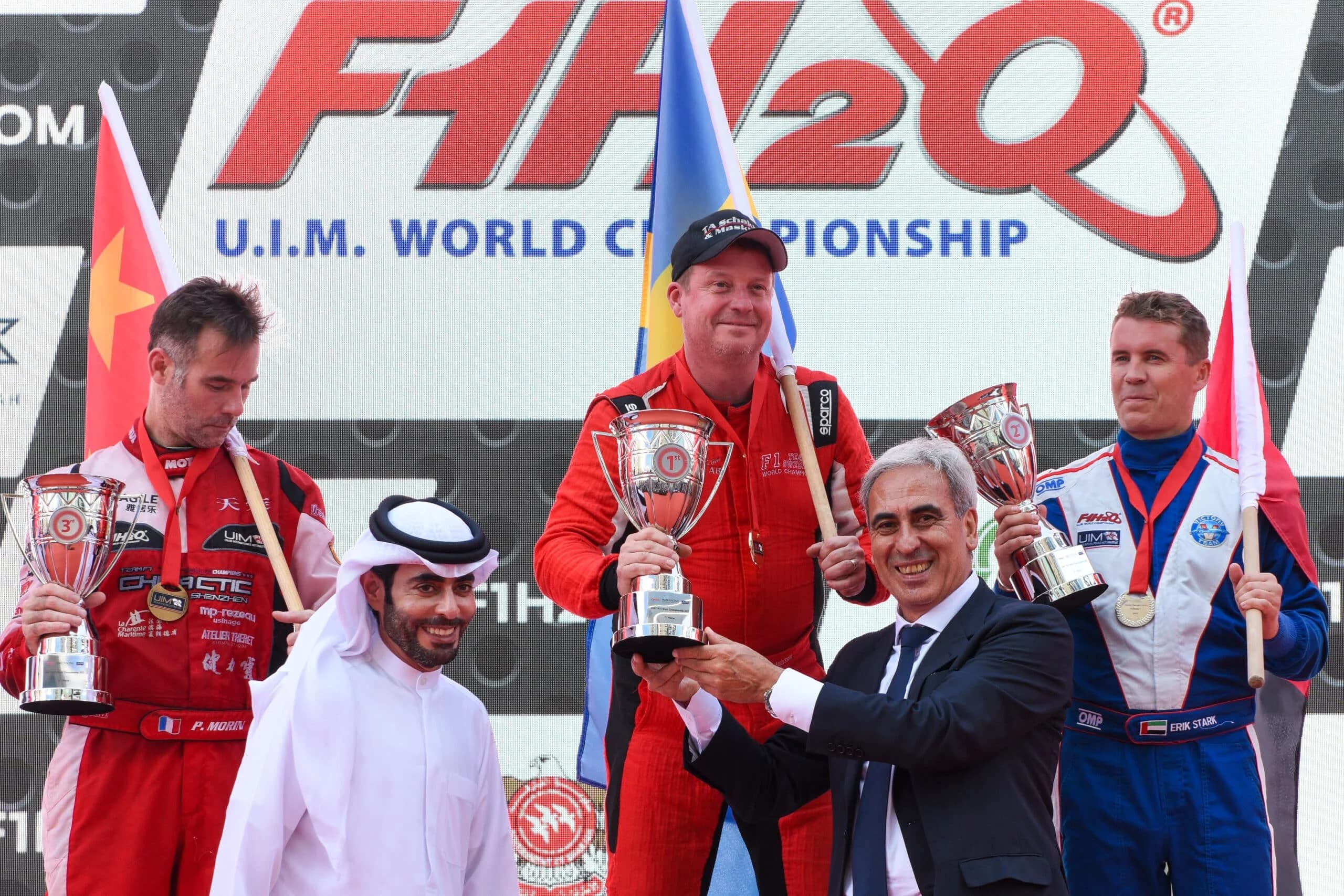Podium ceremony at the F1H2O UIM World Championship with three racers holding trophies while standing on the podium. The first-place winner is holding a trophy and flag, with others standing beside them. Officials in suits are presenting the awards.